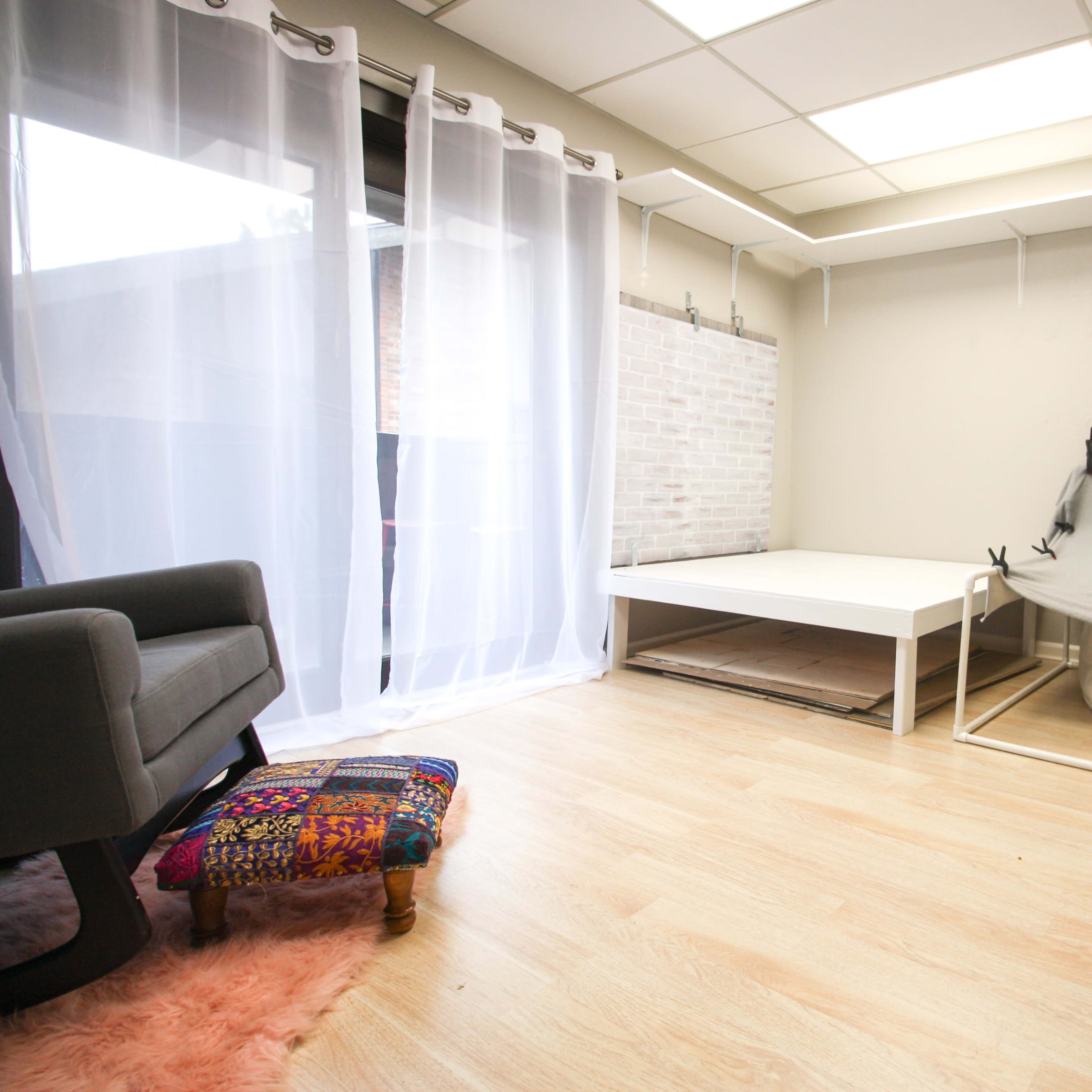 A minimalist room with light wooden flooring, a large window covered by sheer curtains, and a white platform bed alongside a hammock setup.