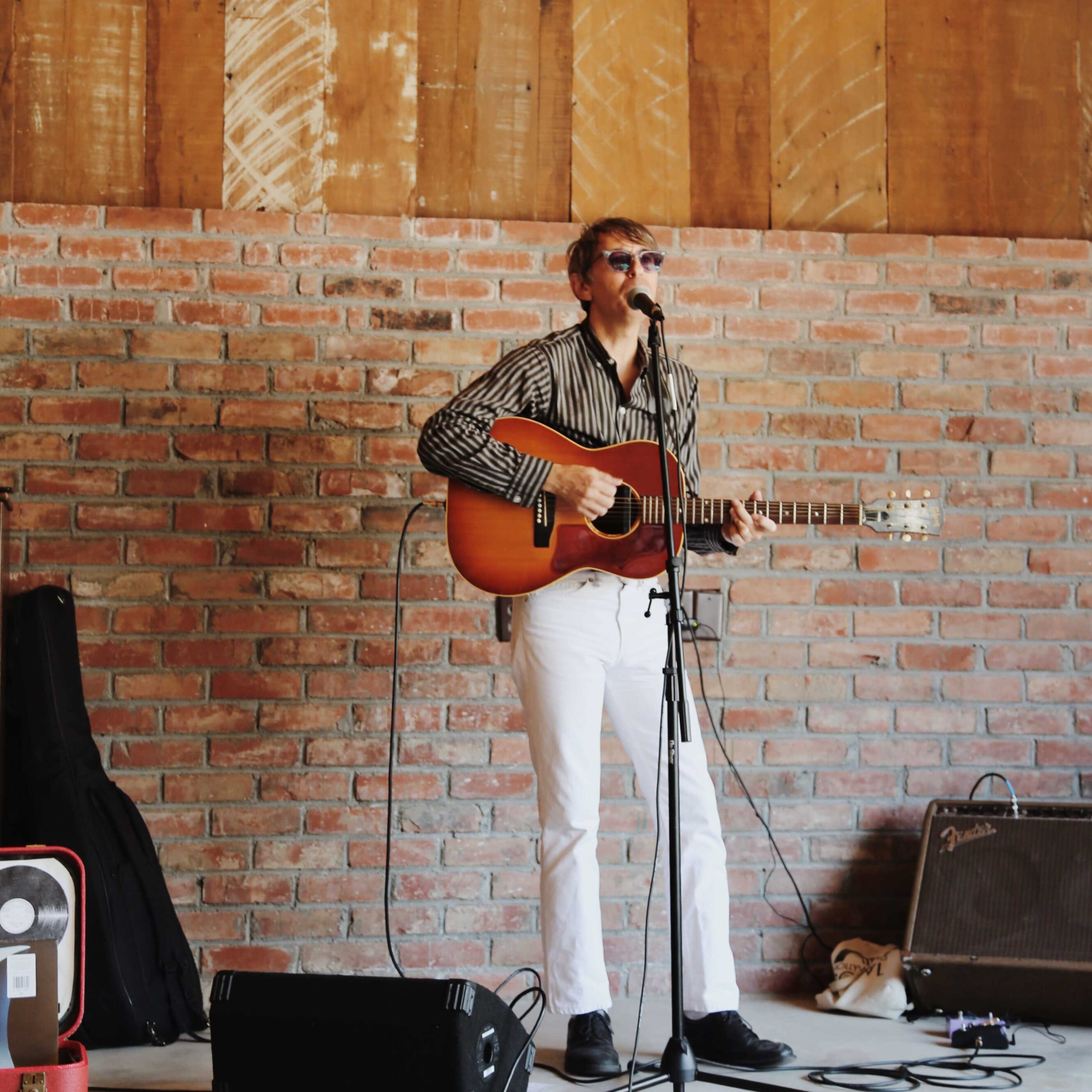 A musician performs on stage with an acoustic guitar in a rustic brick building.
