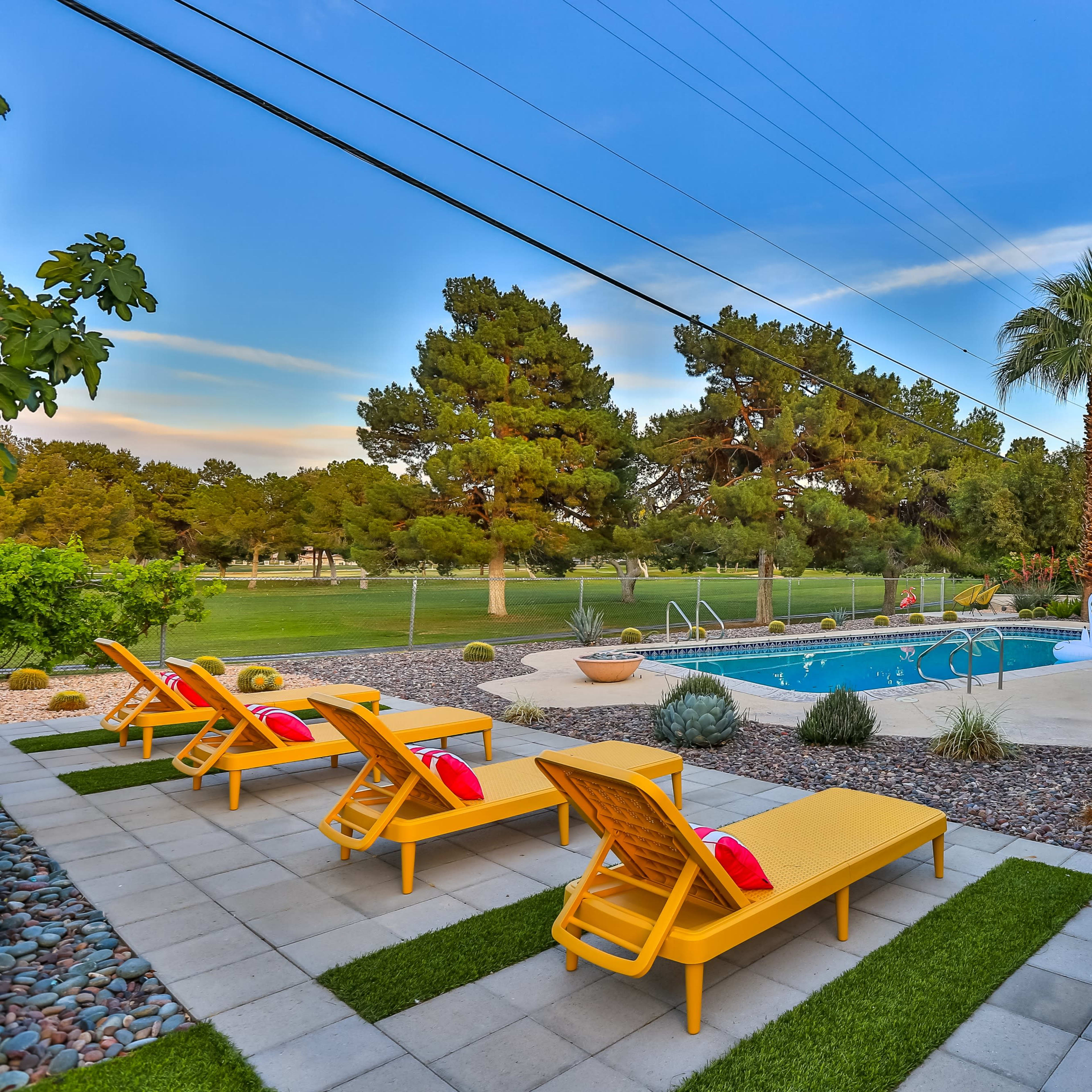 A backyard features a swimming pool surrounded by lounge chairs, palm trees, and landscaped greenery.