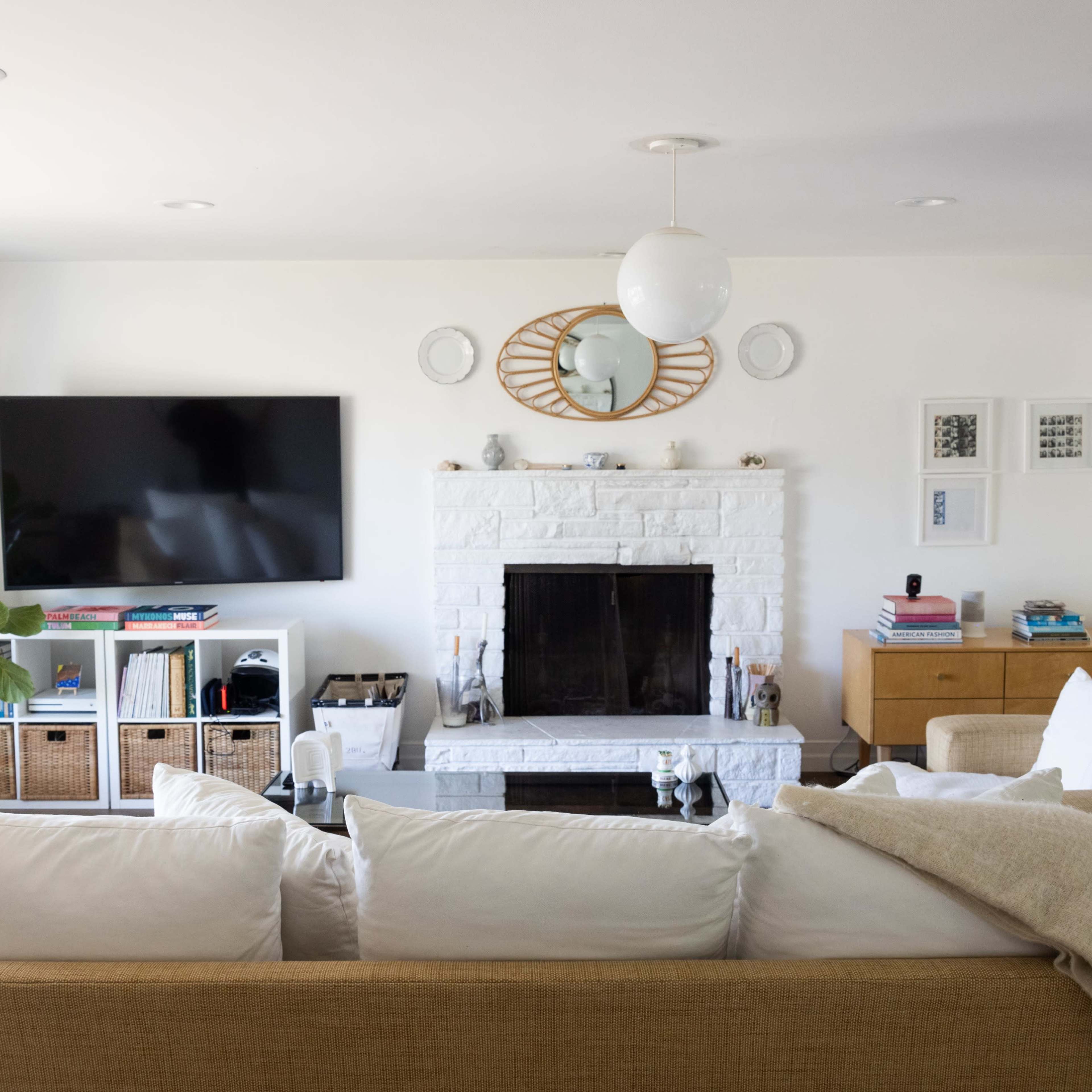 A living room features a white brick fireplace, a mounted TV, a large sectional sofa, and decorative elements including a round mirror and a plant.