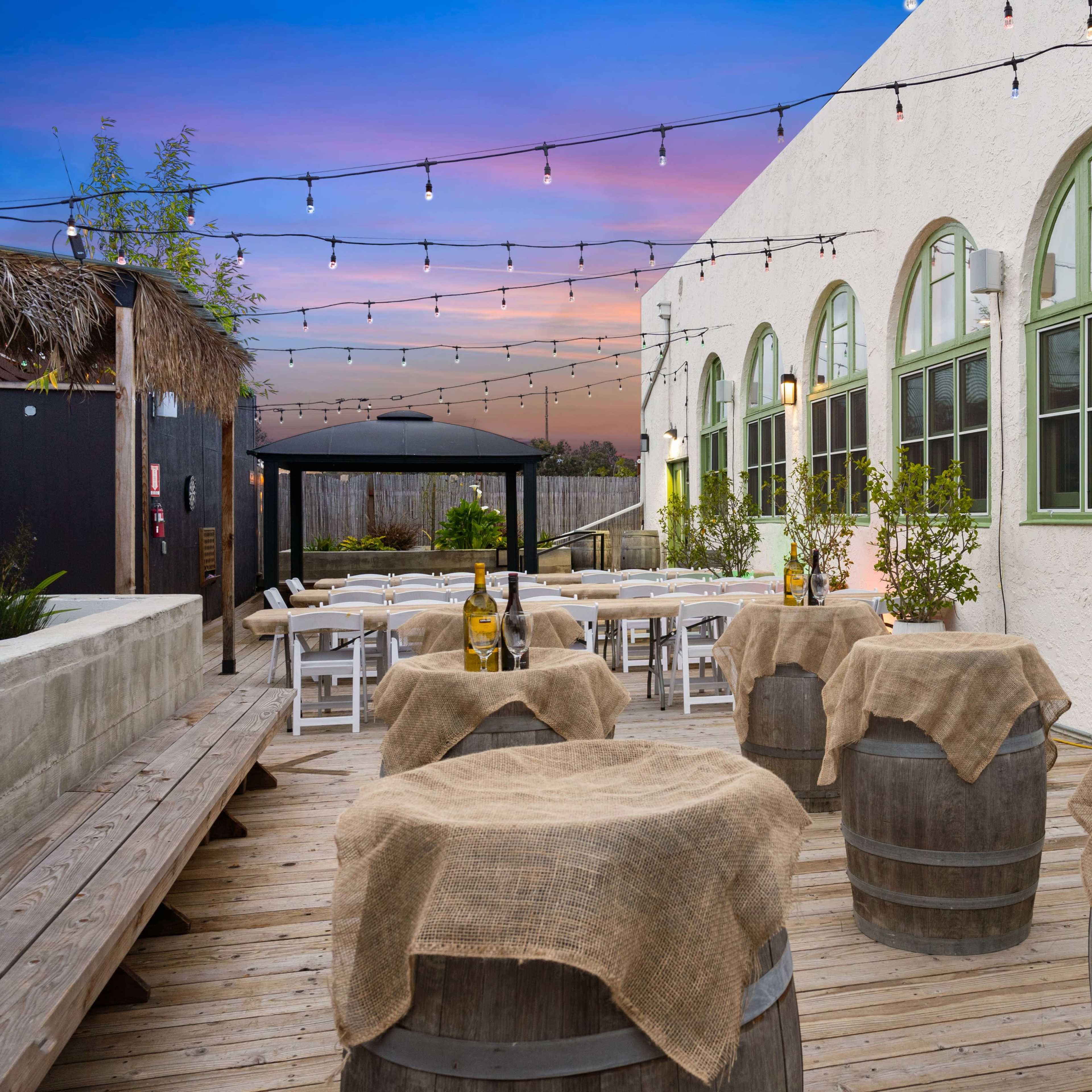 An outdoor patio area with wooden barrels topped with burlap, a barbecue grill, and tables arranged for dining under string lights against a backdrop of a colorful sunset.