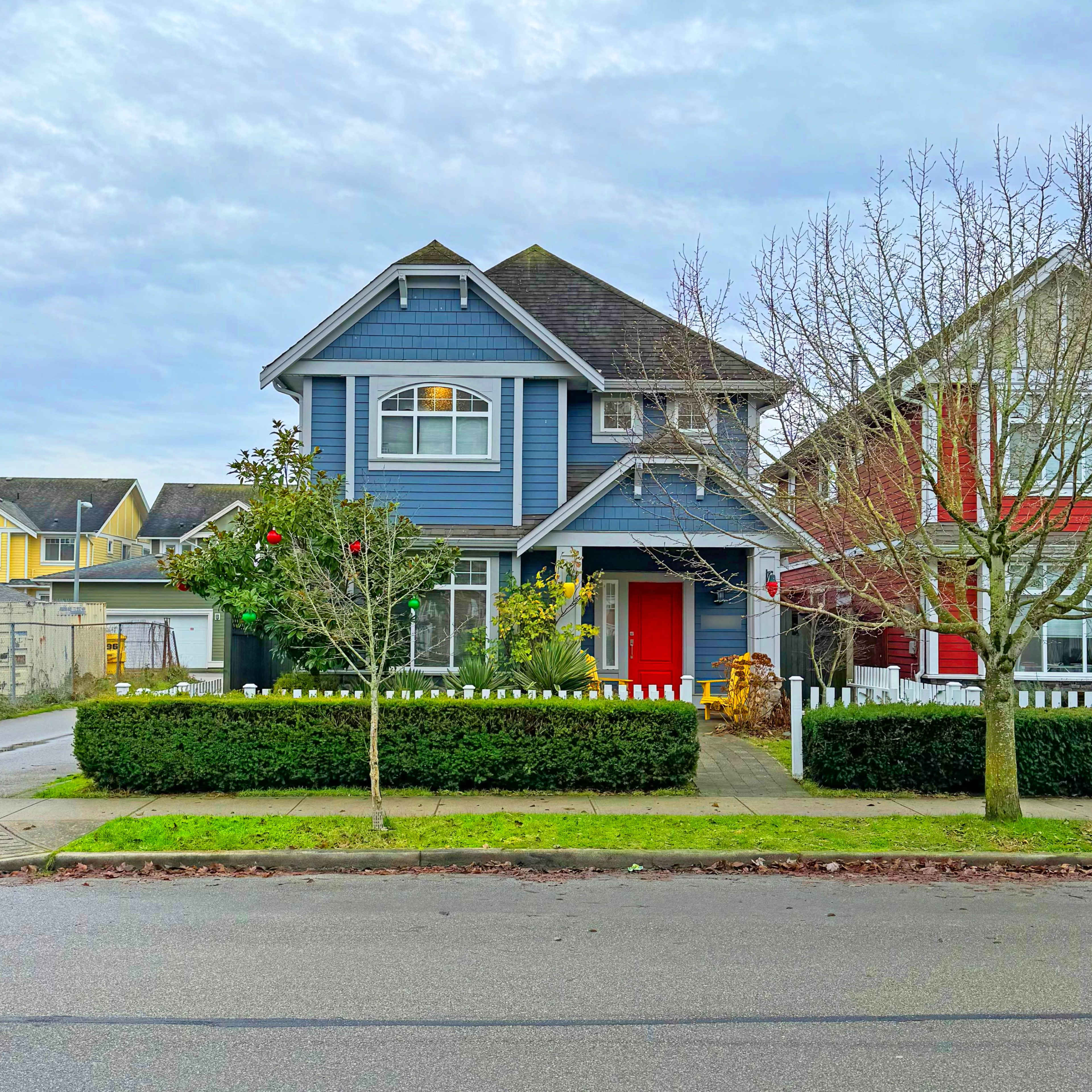 A blue two-story house with a red door is situated on a street lined with trees and a low hedge, alongside a neighboring home.