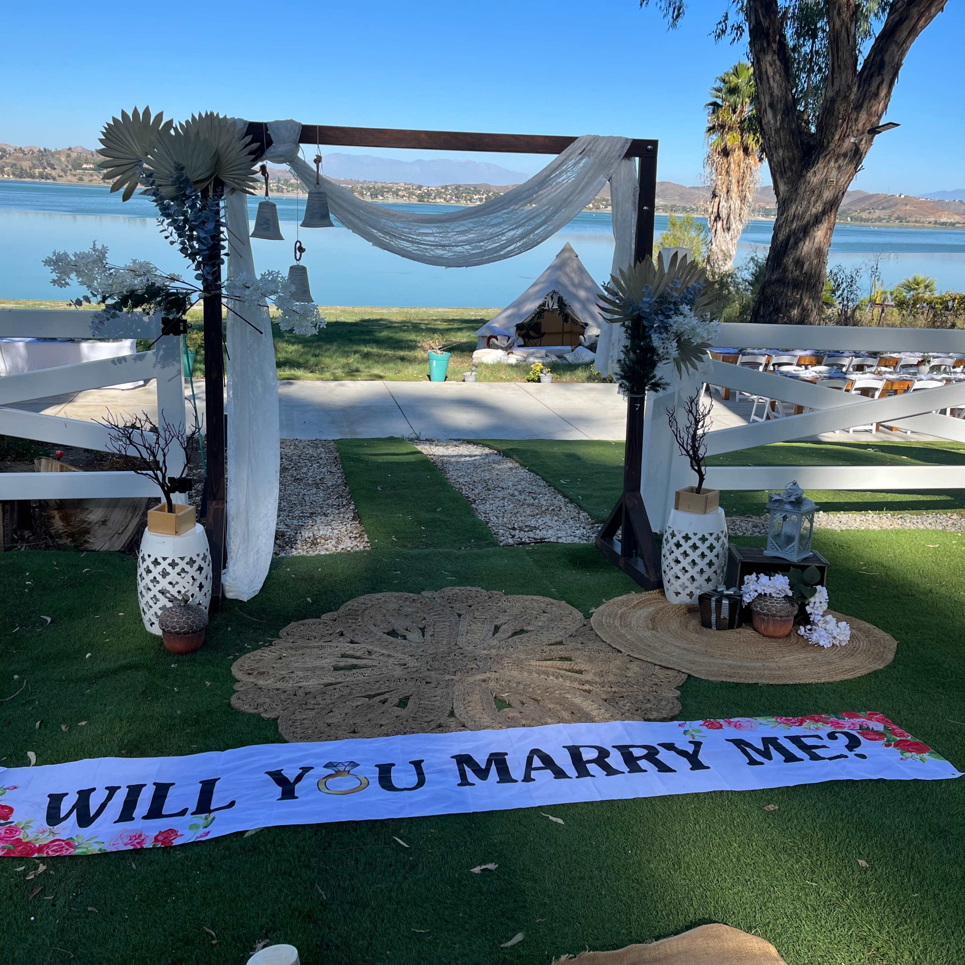The scene features a wedding proposal setup by a lake, adorned with a decorative arch, a large floral banner reading "WILL YOU MARRY ME?", and carefully arranged decorations around a green lawn.