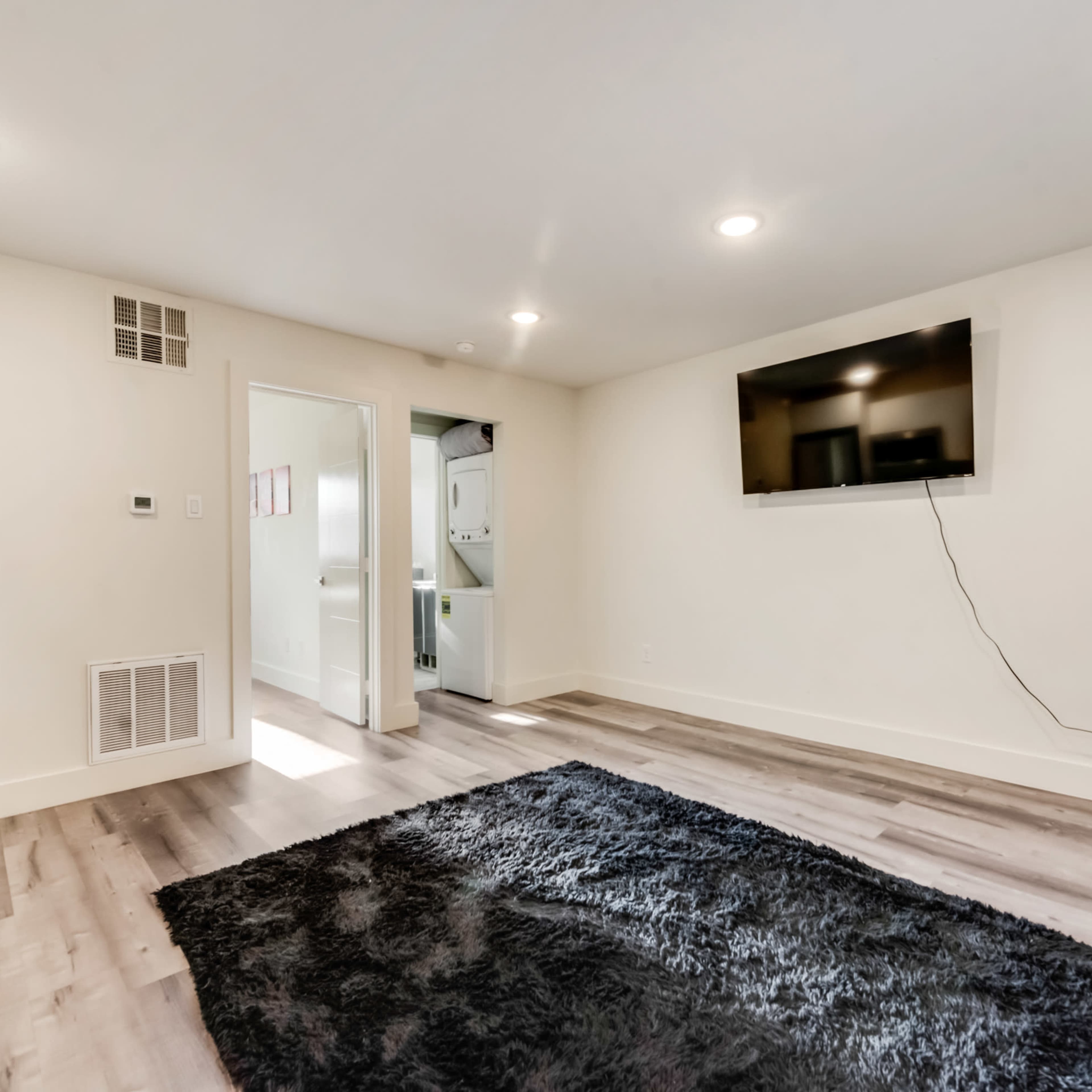 The room features light-colored walls, a flat-screen TV mounted on the wall, and a dark rug on the floor, with a doorway leading to another area.