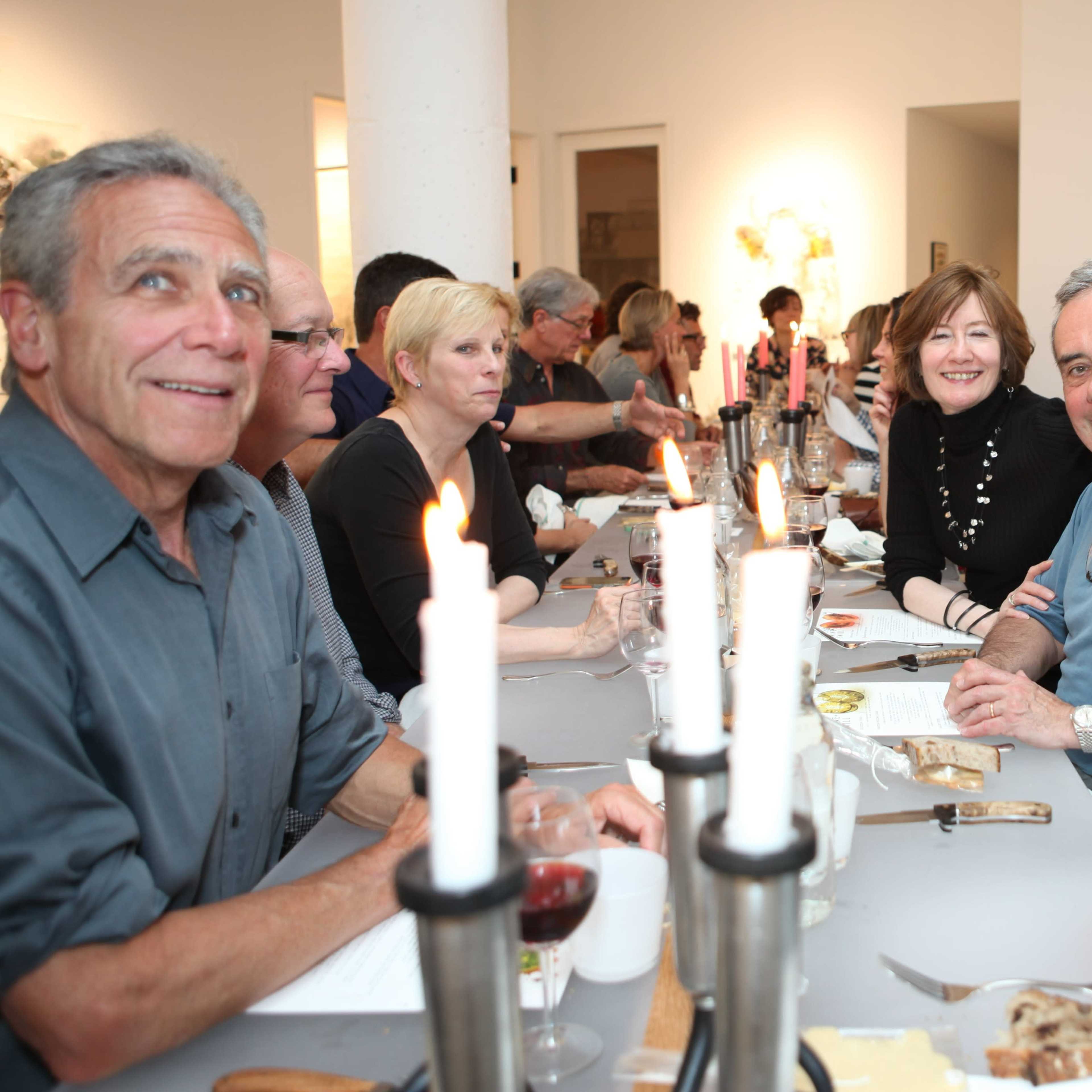 A group of seven people sits around a well-set dinner table, with lit candles in the foreground and artworks visible on the walls.