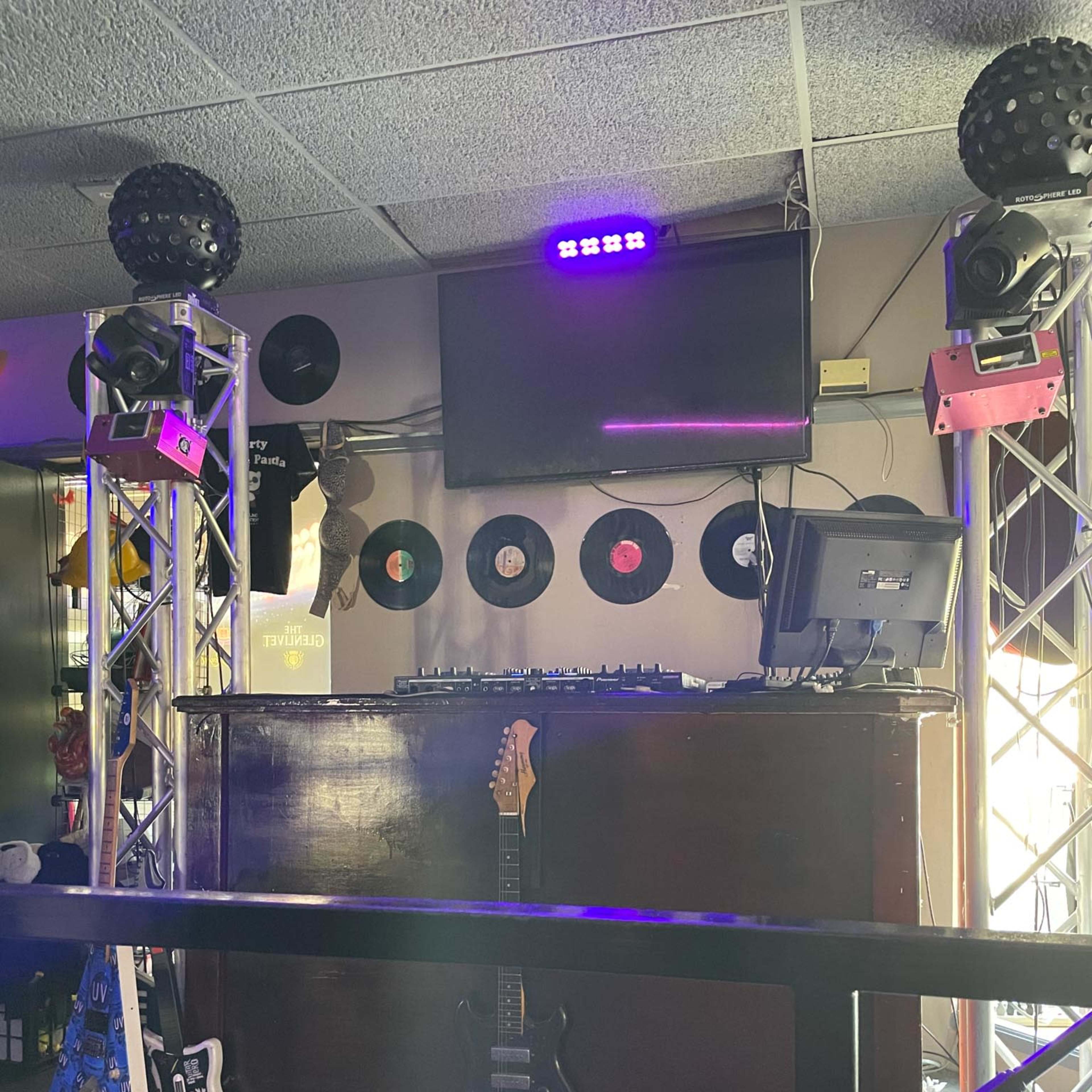 A DJ booth with turntables, a computer monitor, and vinyl records mounted on the wall, illuminated by colorful stage lights.