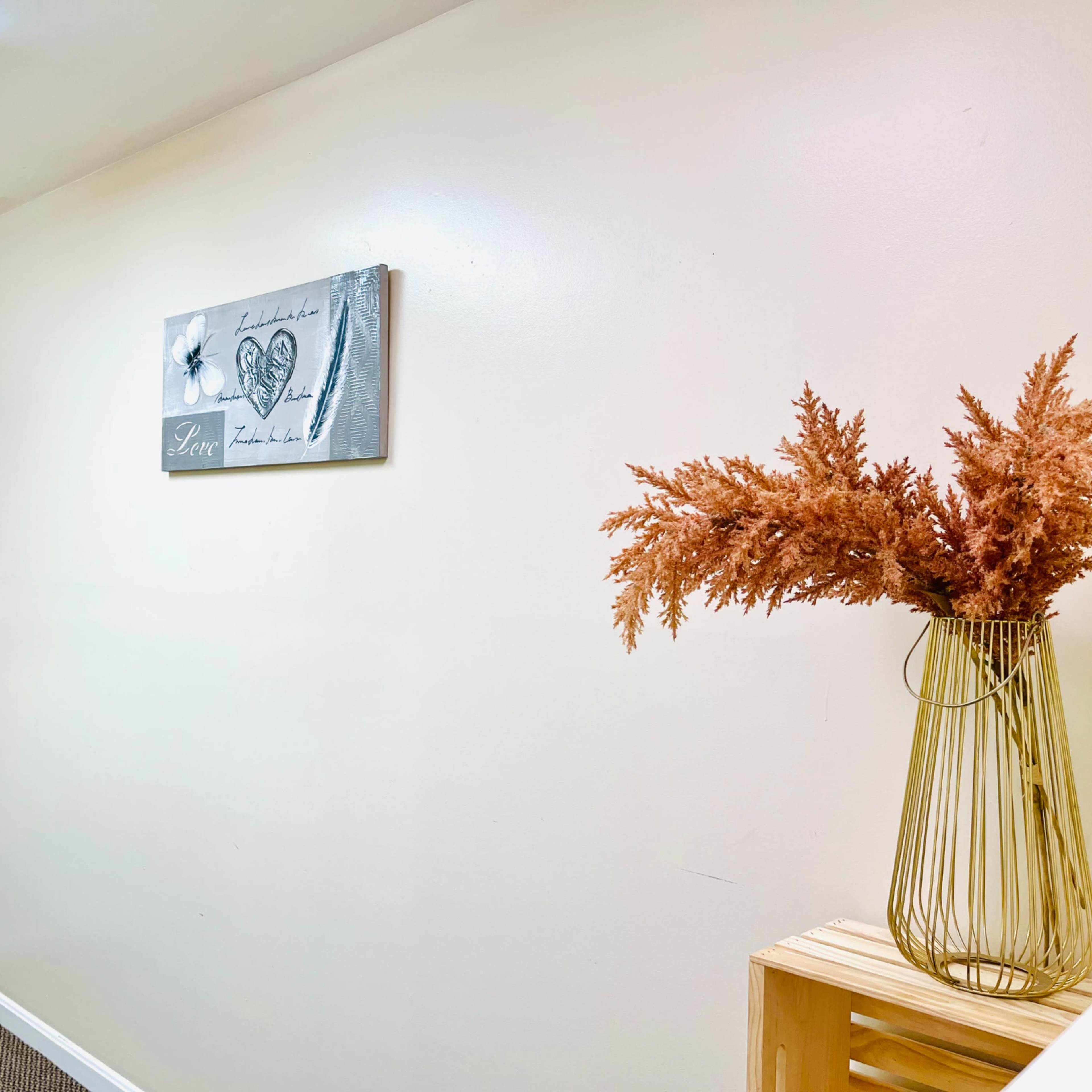 A small, simple room with a table, a wall decoration featuring a figure in traditional attire, and a vase of dried orange flowers.