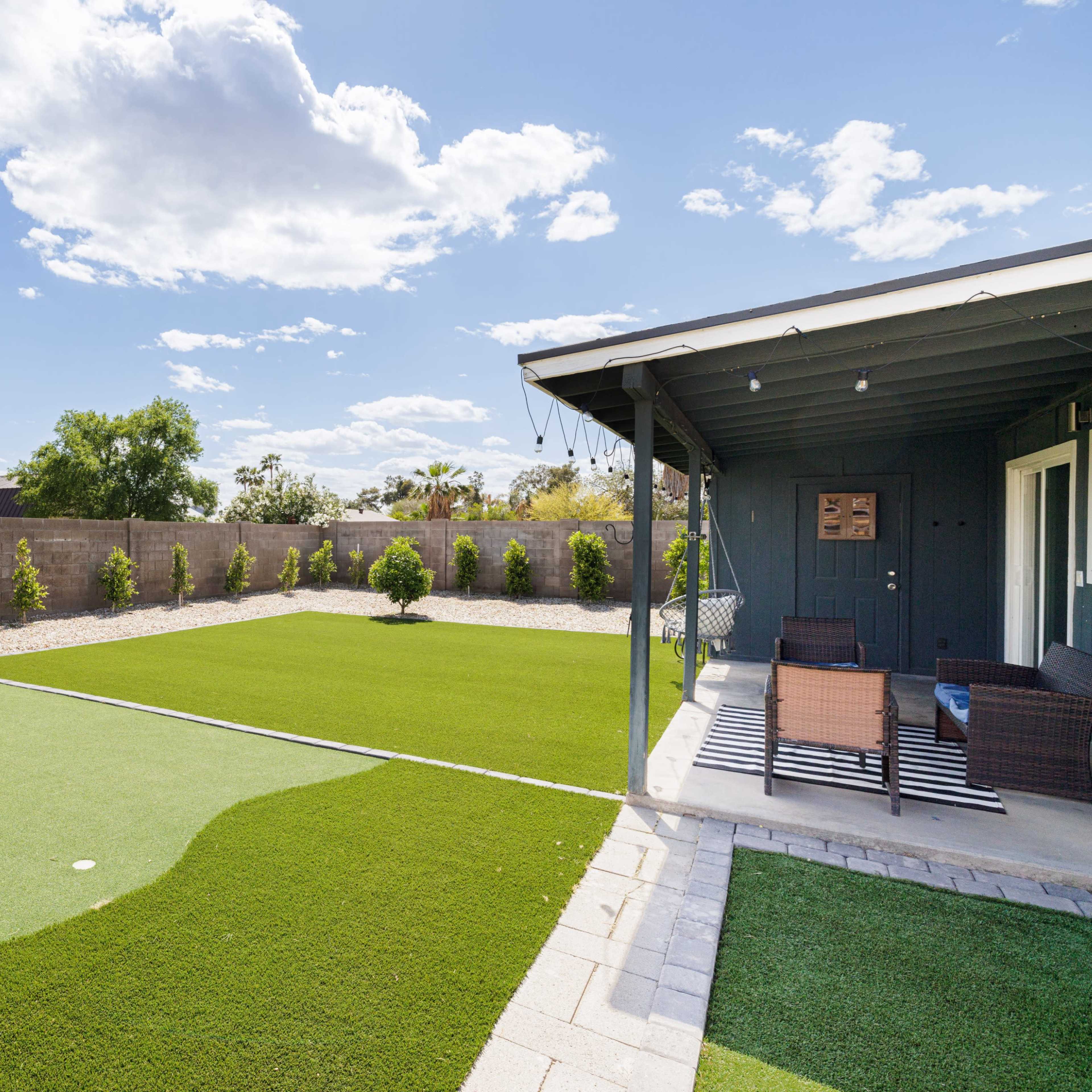 A landscaped backyard with artificial turf, bordered by low shrubs and a patio area with seating.