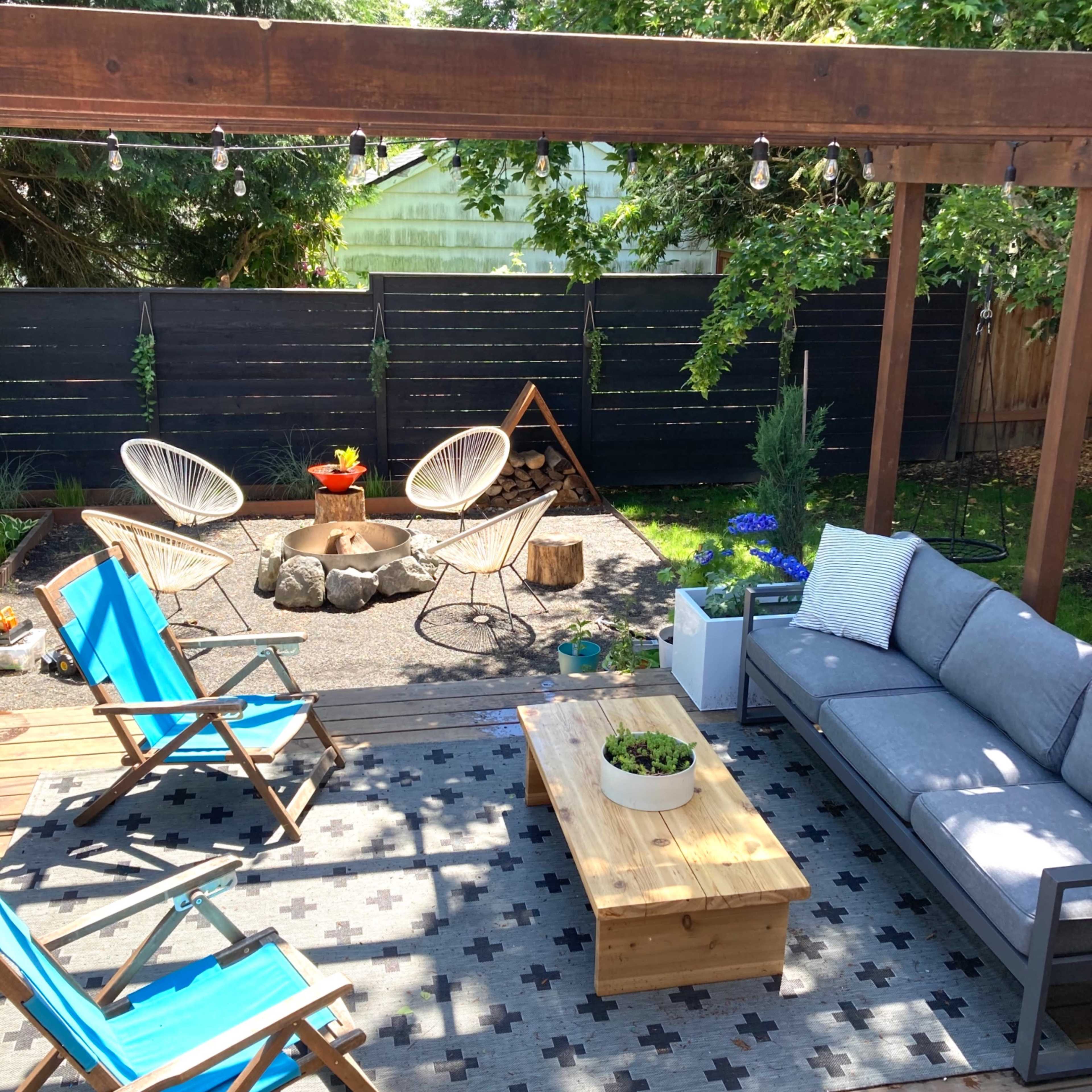 A backyard patio area with outdoor seating, a wooden coffee table, and garden decorations surrounded by greenery.