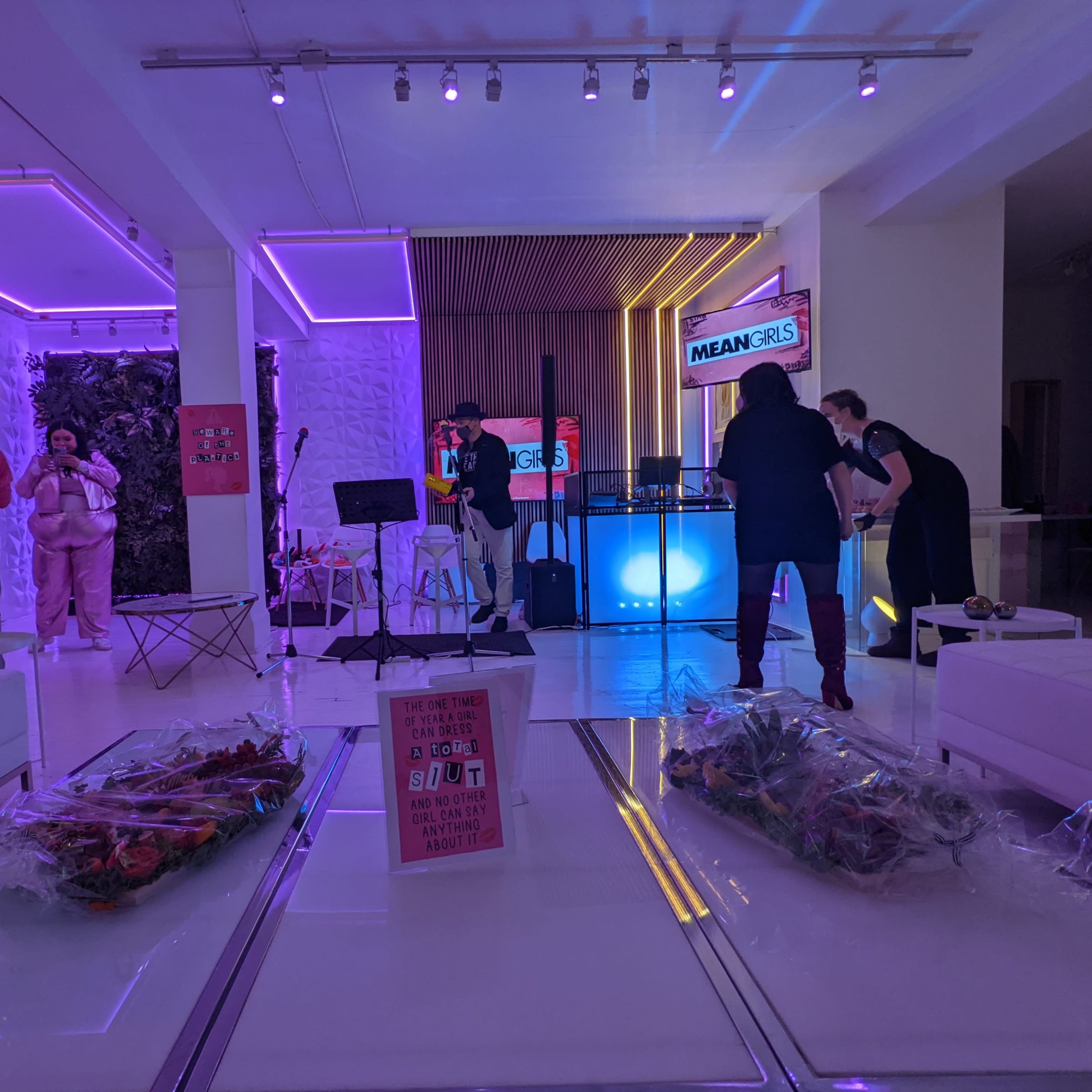 The scene shows a brightly lit event space with purple lighting, featuring a stage area and attendees preparing for a performance or gathering.