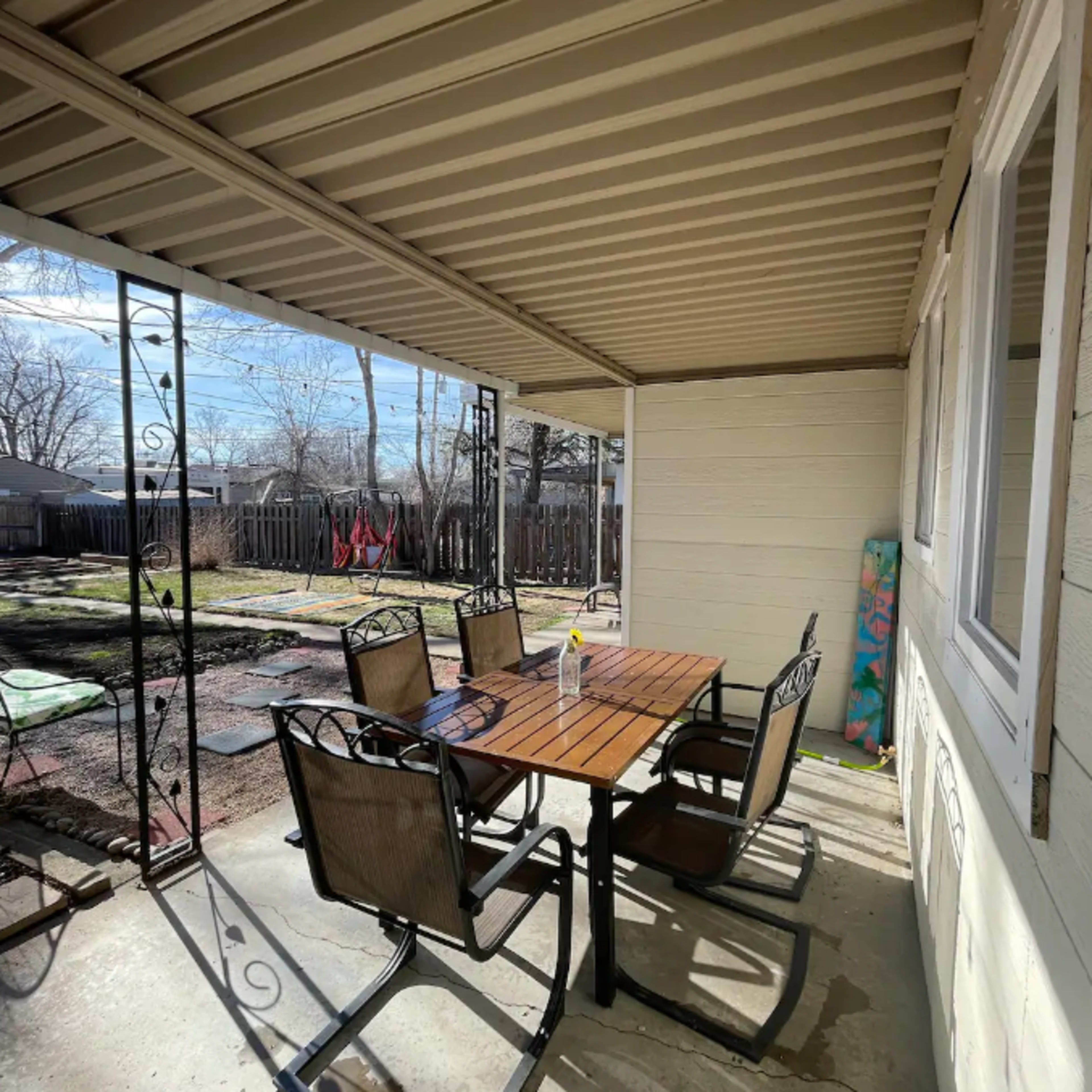 A patio area features a wooden table surrounded by six metal chairs, with a view of a yard in the background.
