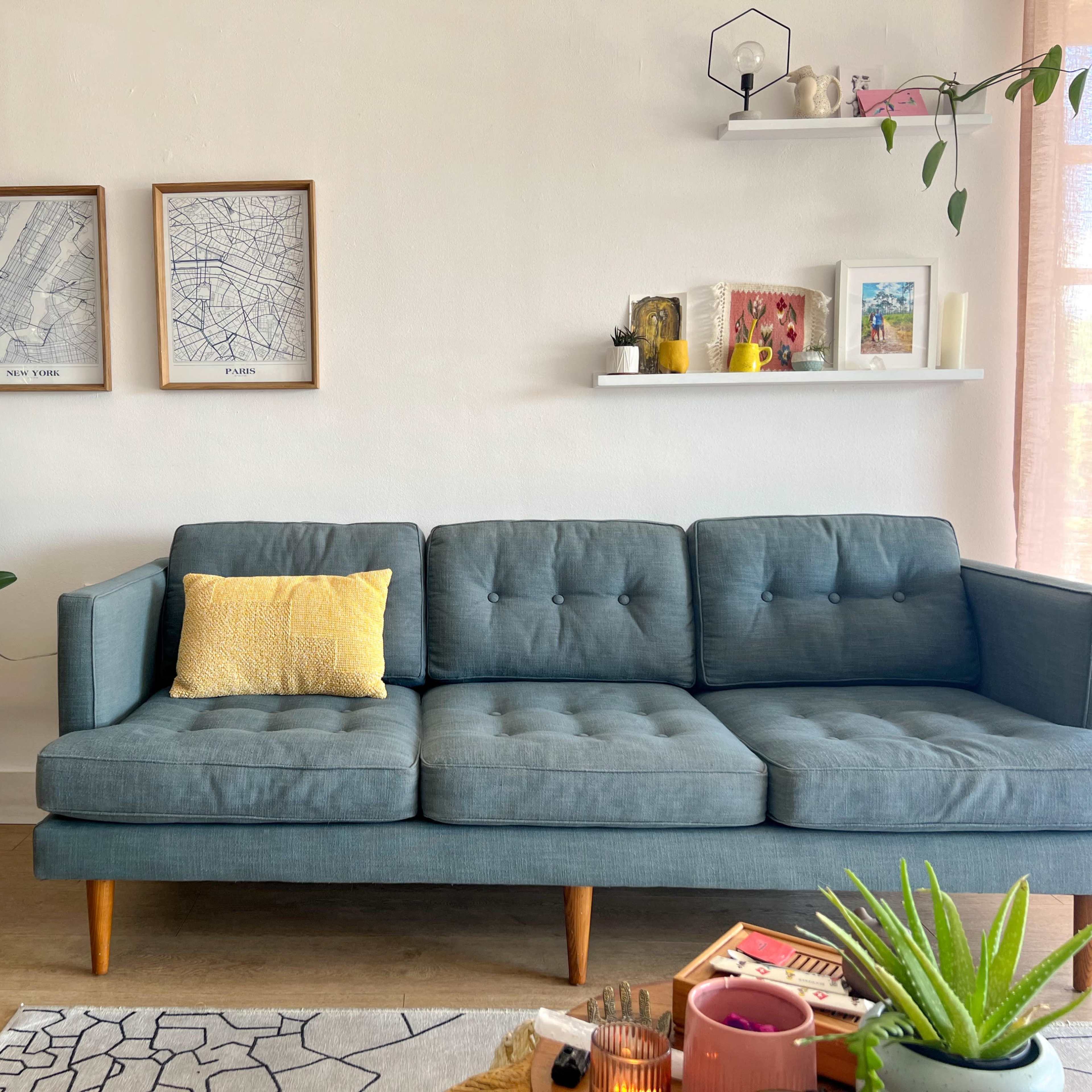 A modern living room features a teal sofa with a yellow cushion, a round side table with plants and a lamp, framed maps on the wall, and shelves displaying decorative items.
