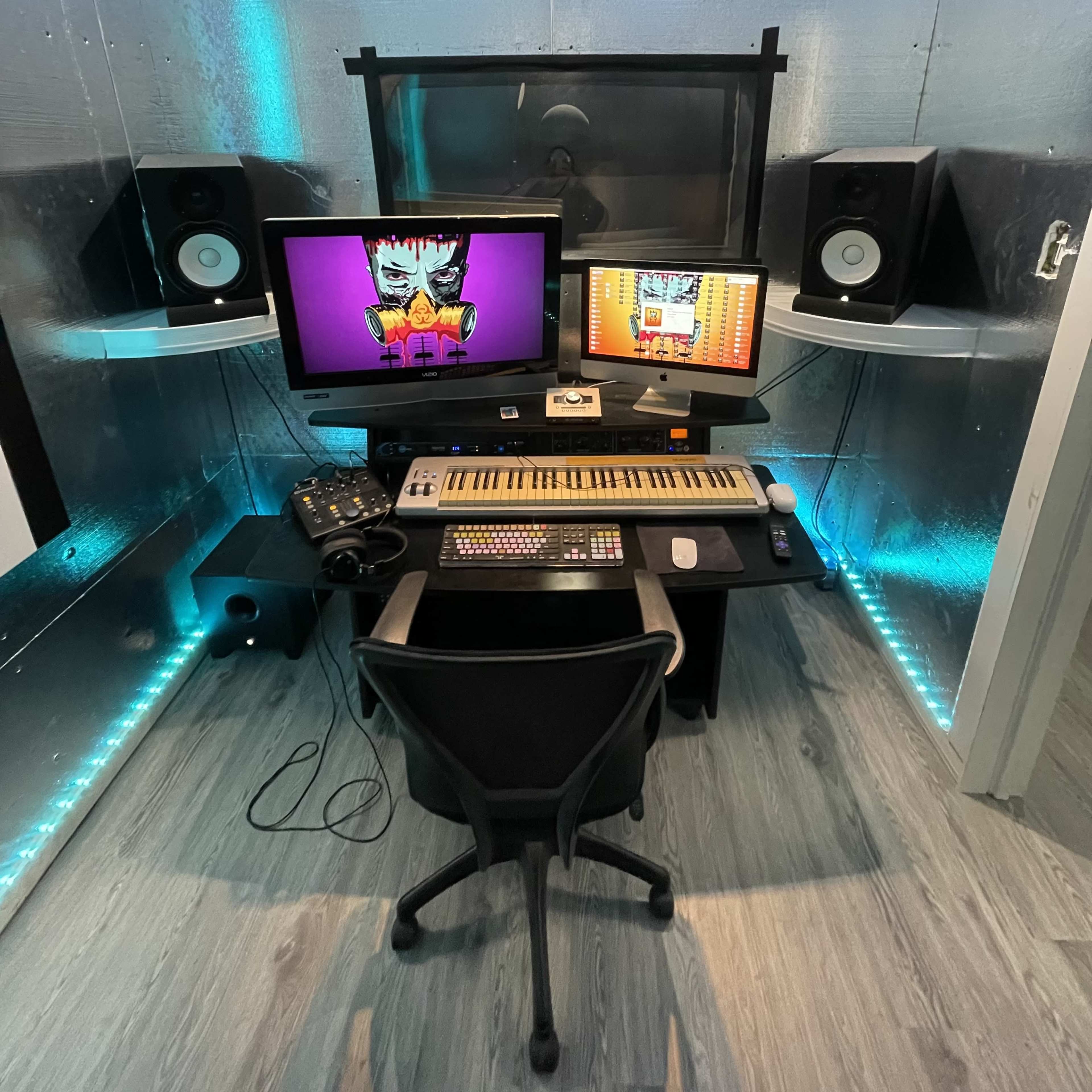 A well-equipped music production studio features a keyboard, multiple monitors, speakers, and a computer, all set against a metallic wall with ambient lighting.