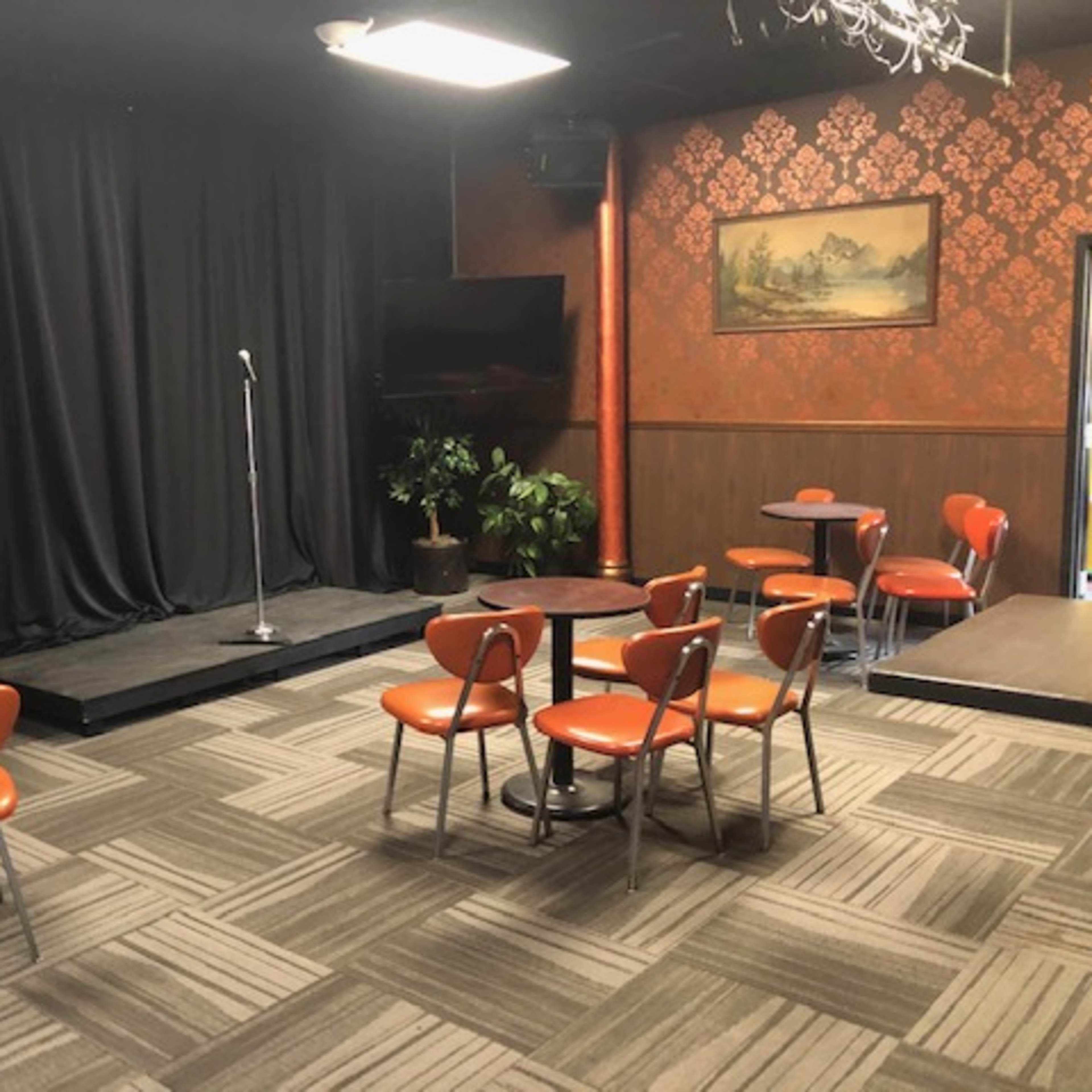 A small performance space with a black curtain, a microphone stand on a stage, and several round tables with orange chairs arranged around the room.