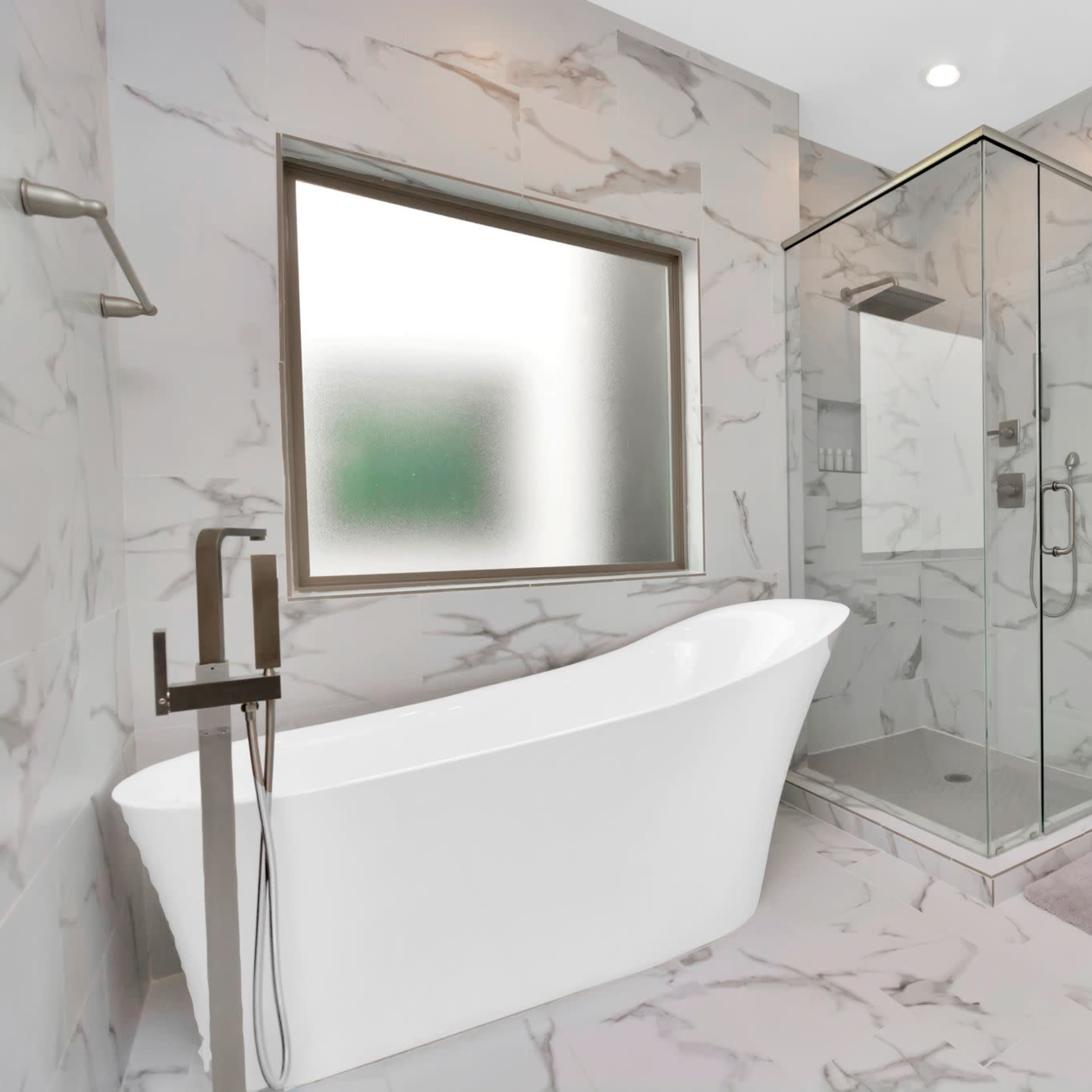 A modern bathroom featuring a freestanding white bathtub, a glass shower enclosure, and marble-patterned walls and flooring.