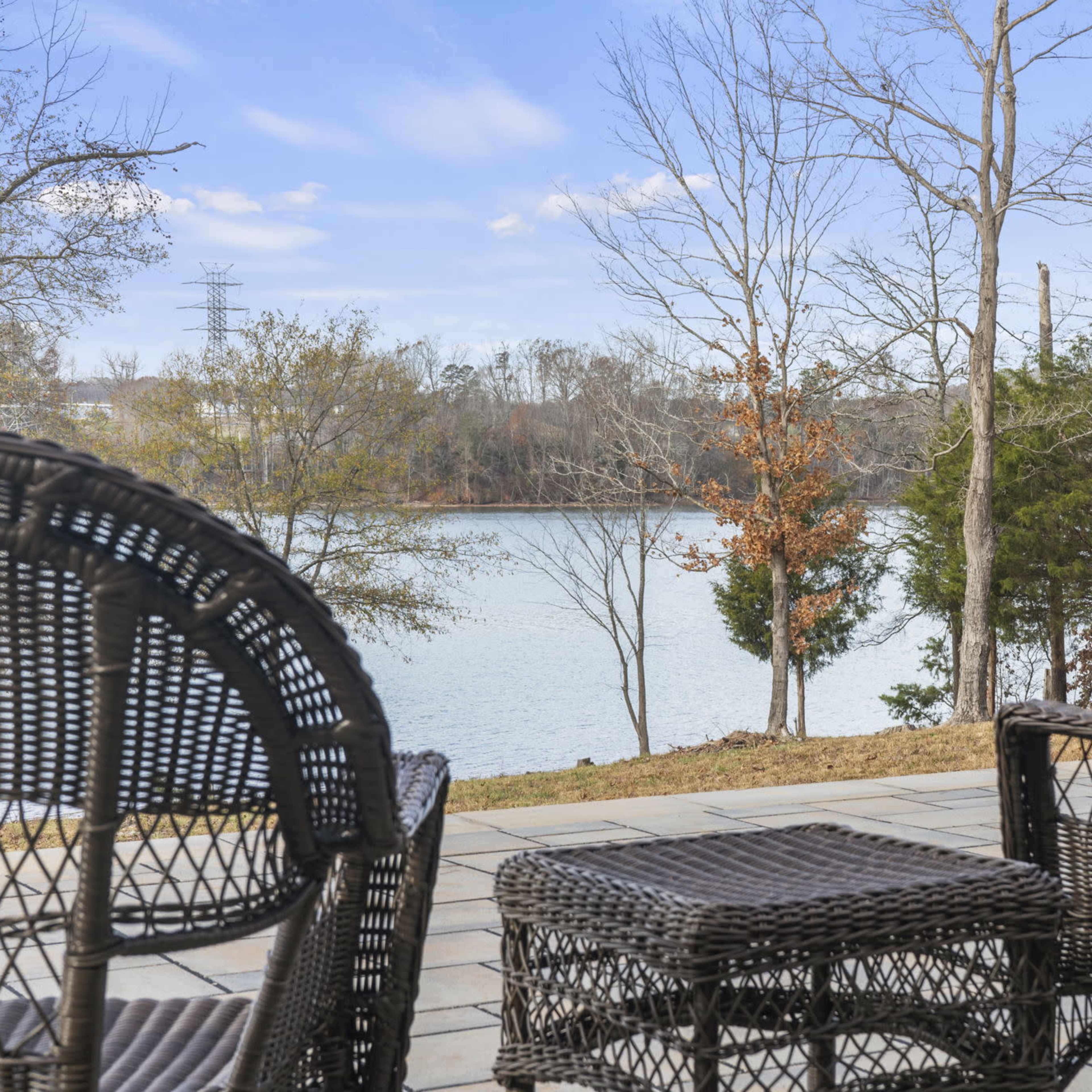 A patio area with wicker chairs overlooks a calm lake surrounded by trees.