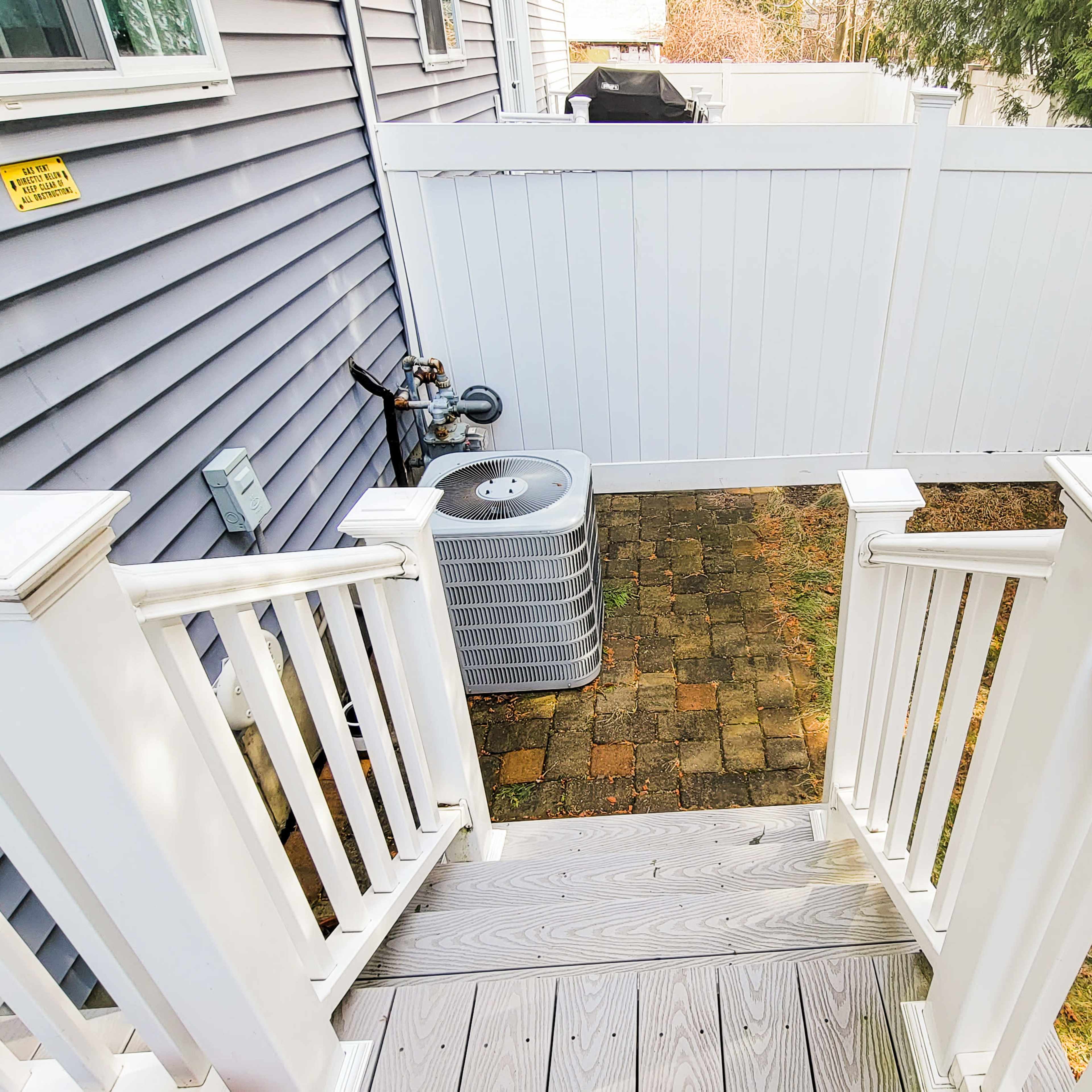 A set of wooden steps leading down from a house to a small, fenced backyard with pavers and an air conditioning unit.