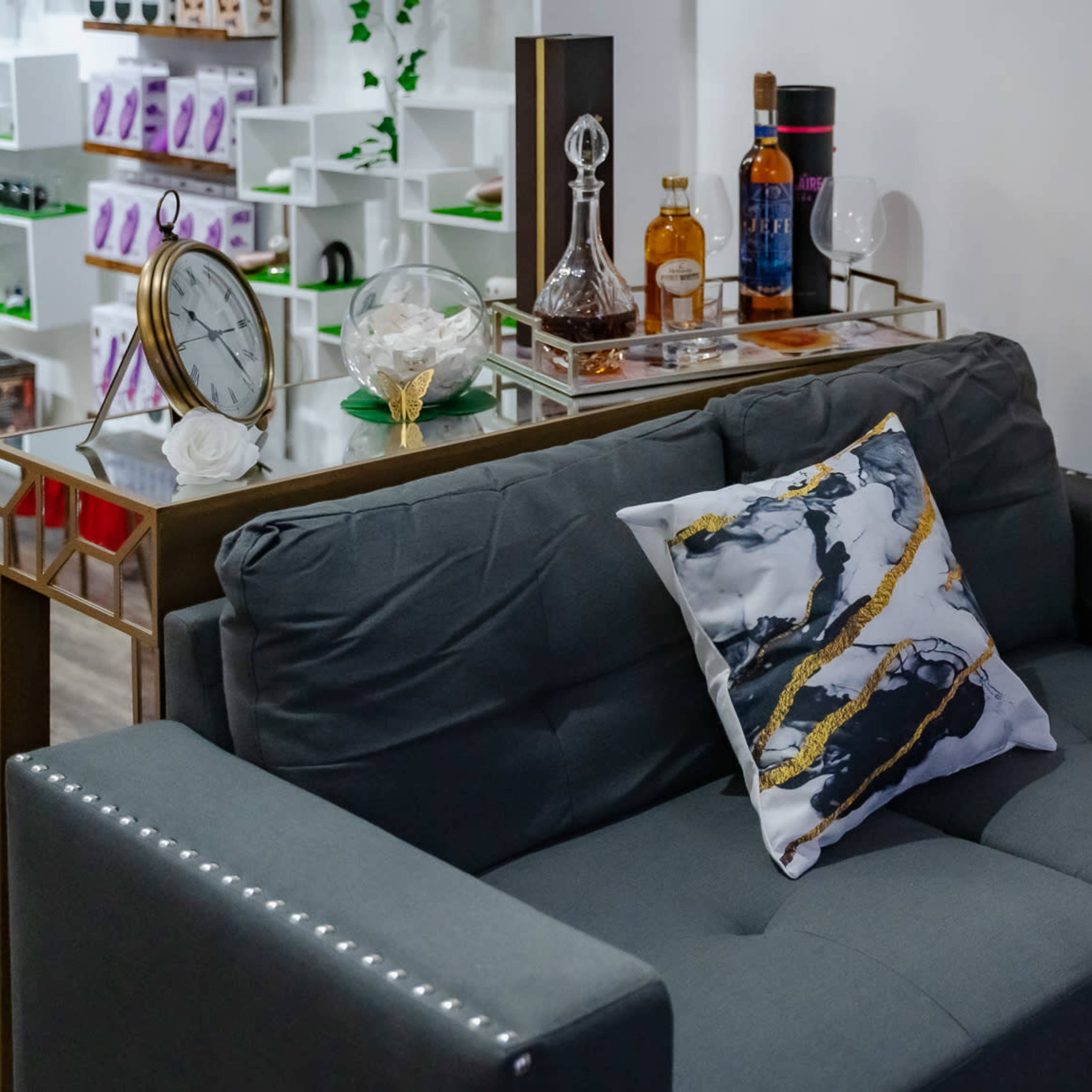 A gray couch with a decorative pillow is placed beside a small table holding a lamp and a sign, with a bar cart in the background displaying various bottles and decorative items.