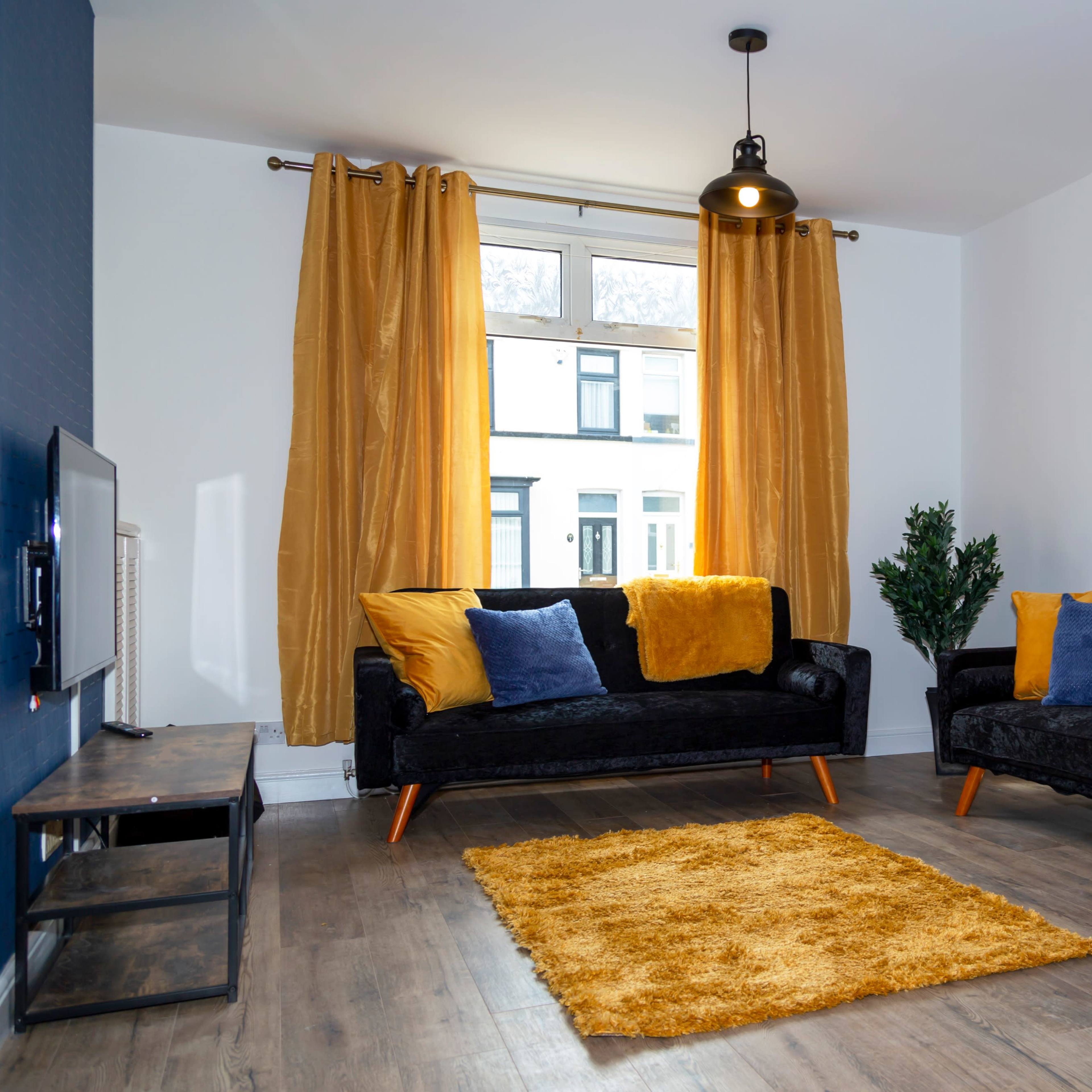 A modern living room features two black sofas with yellow and blue pillows, a yellow rug, and large windows dressed with mustard curtains.