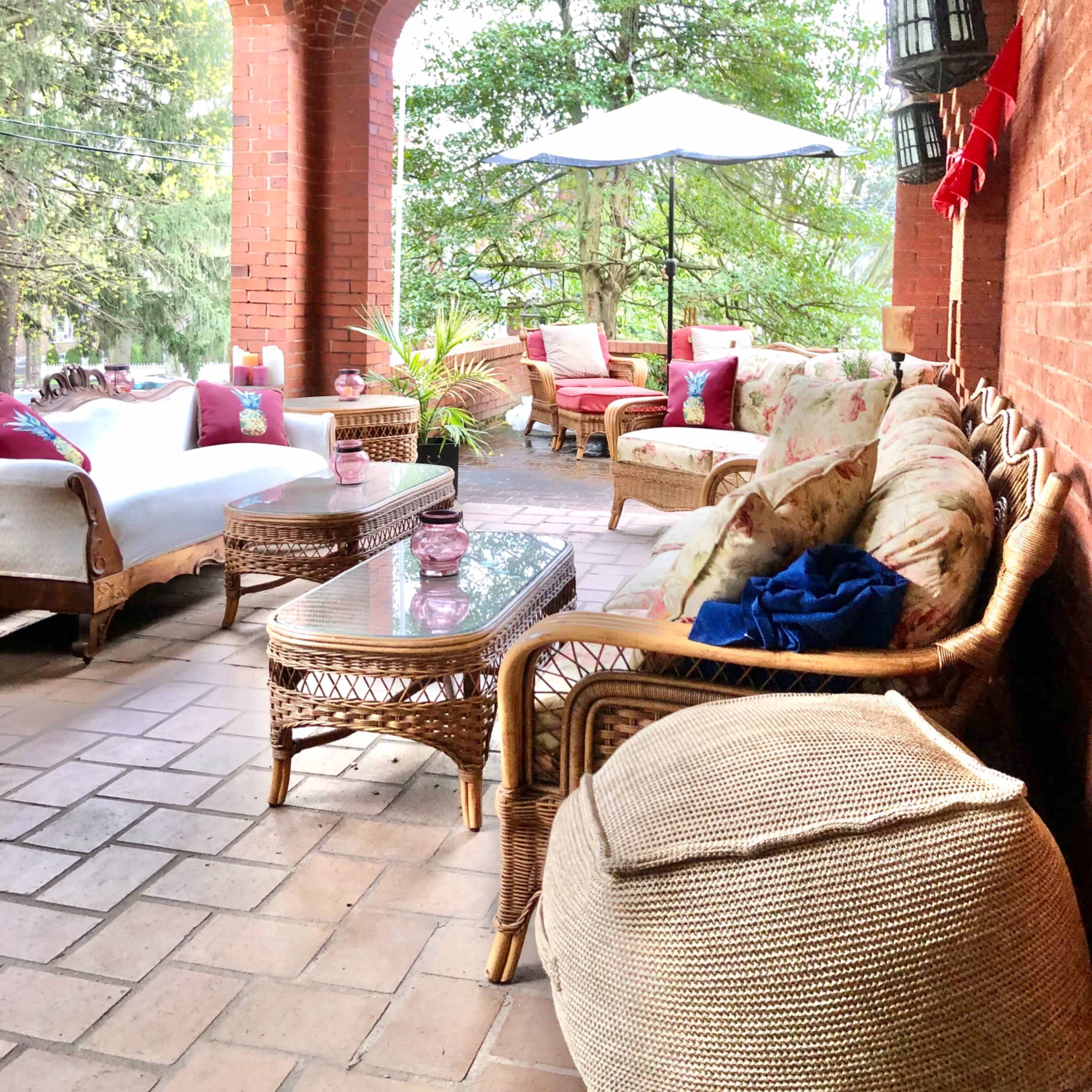 A covered outdoor patio with wicker furniture, including a sofa and armchairs, arranged around a coffee table under a large umbrella.