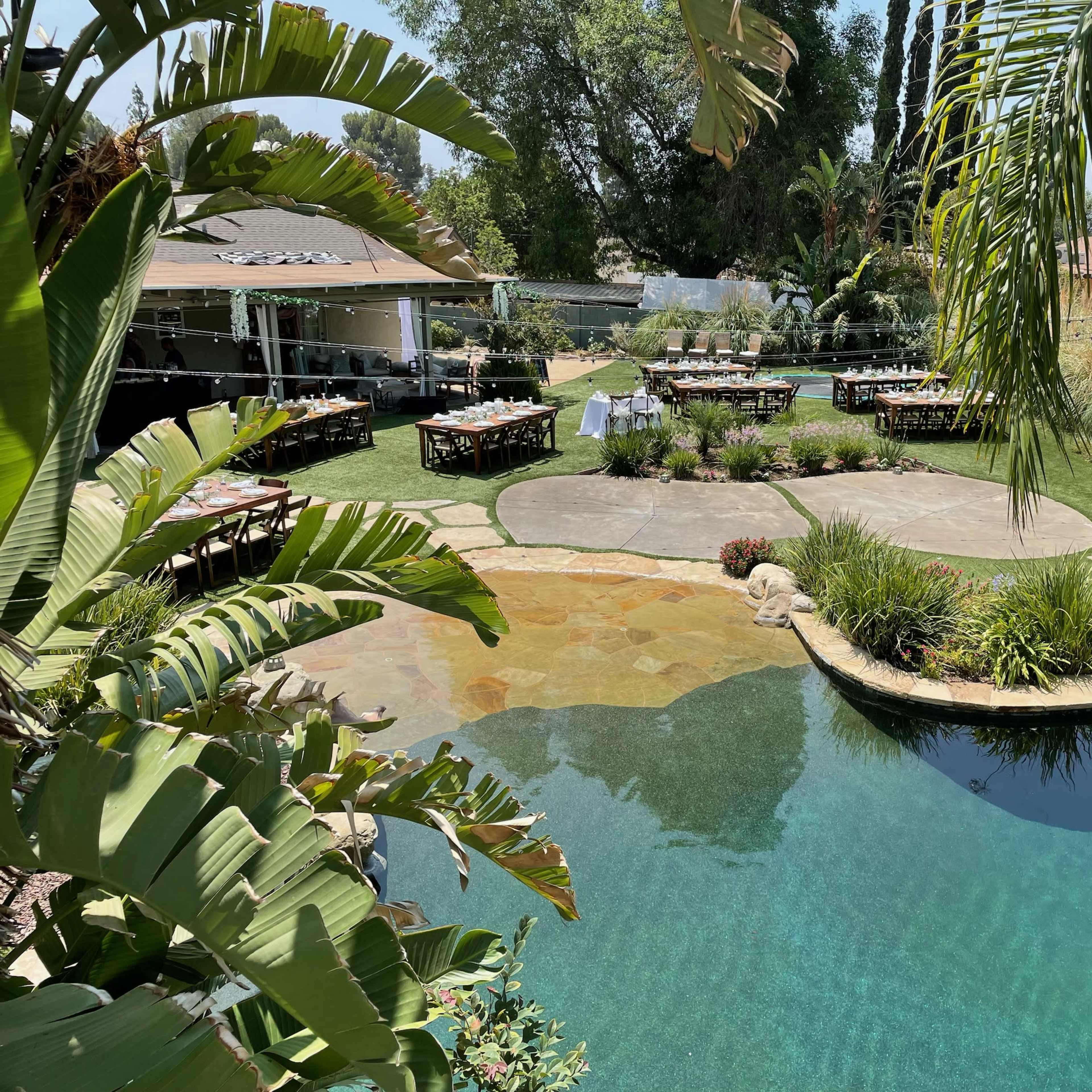A landscaped backyard featuring a swimming pool, stone pathways, and multiple dining tables set up for an outdoor gathering.