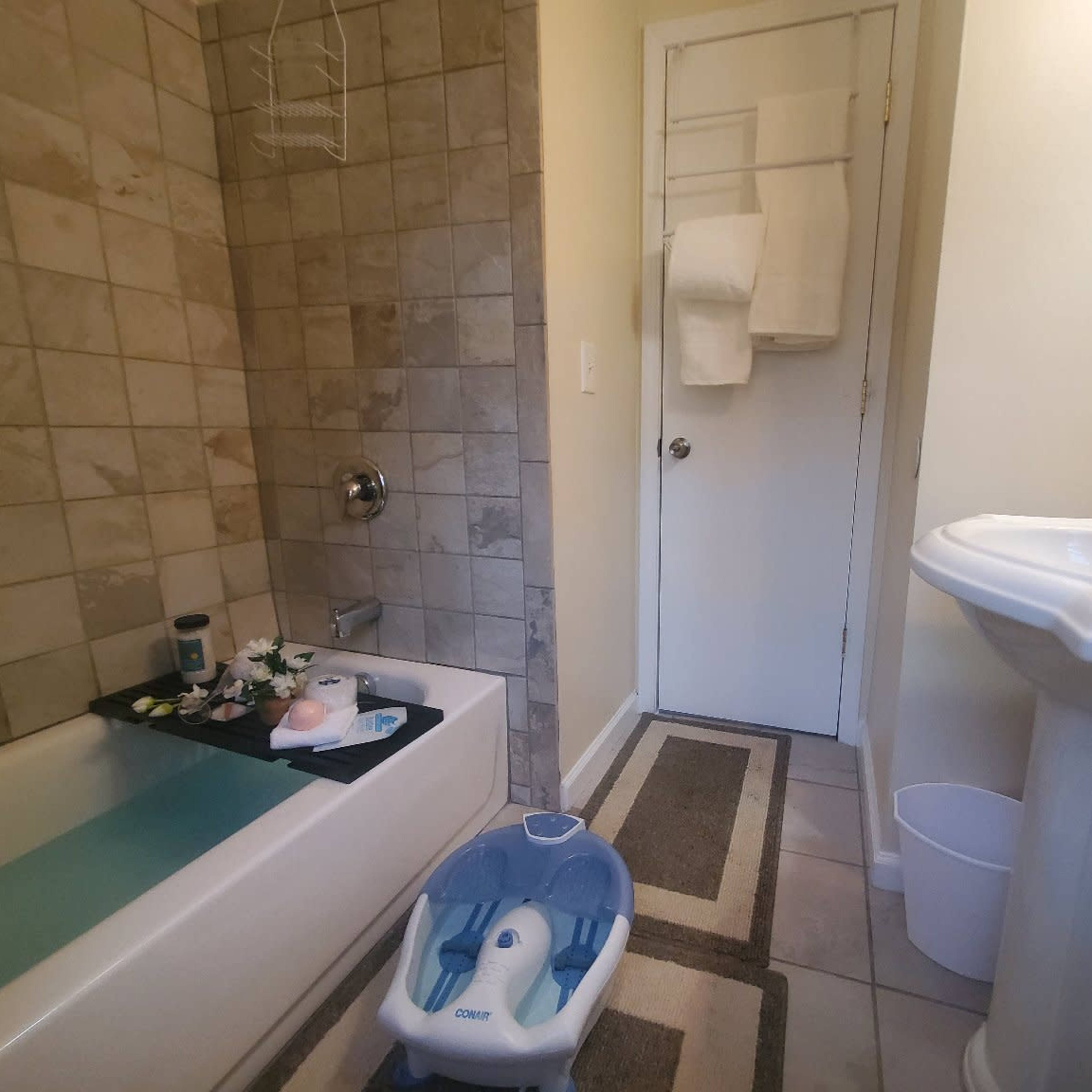 A bathroom with a bathtub, a sink, and a closed door, featuring tiled walls and a bath mat on the floor.