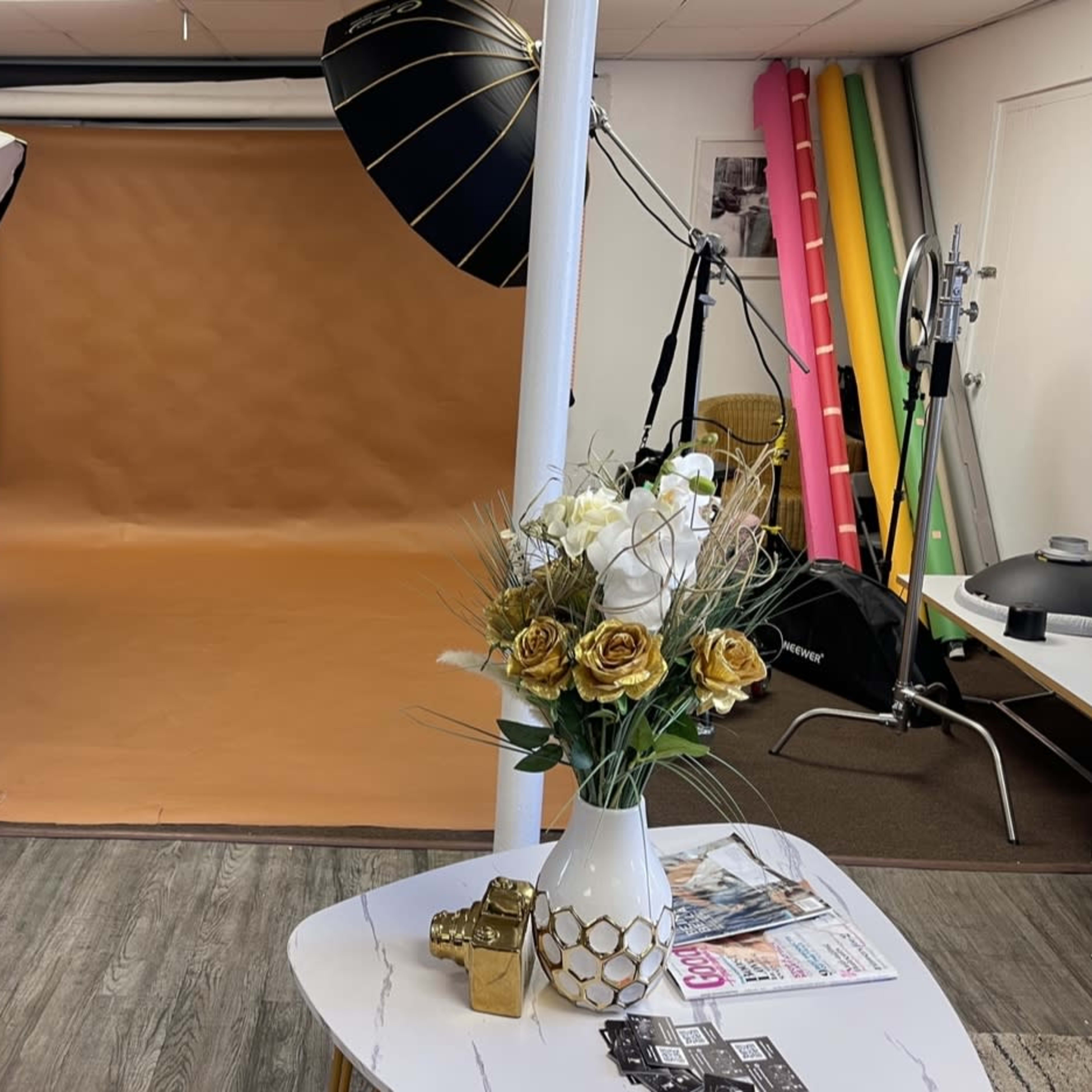 A photography studio featuring a brown backdrop, two large softbox lights, a decorative flower arrangement on a table, and various photography equipment in the background.