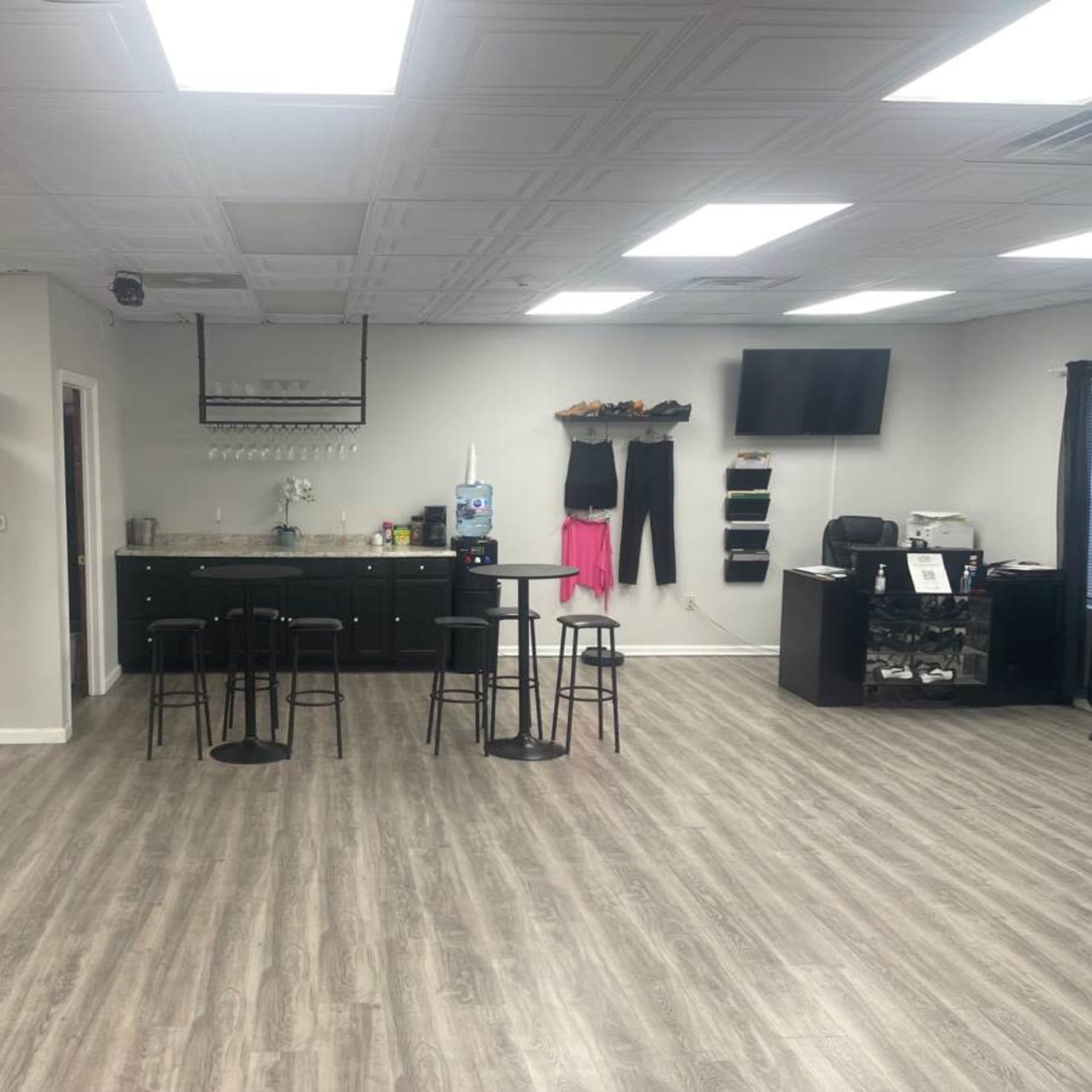 A spacious room featuring a kitchenette area with bar stools, a black shelving unit, and office furniture against a light-colored wall.