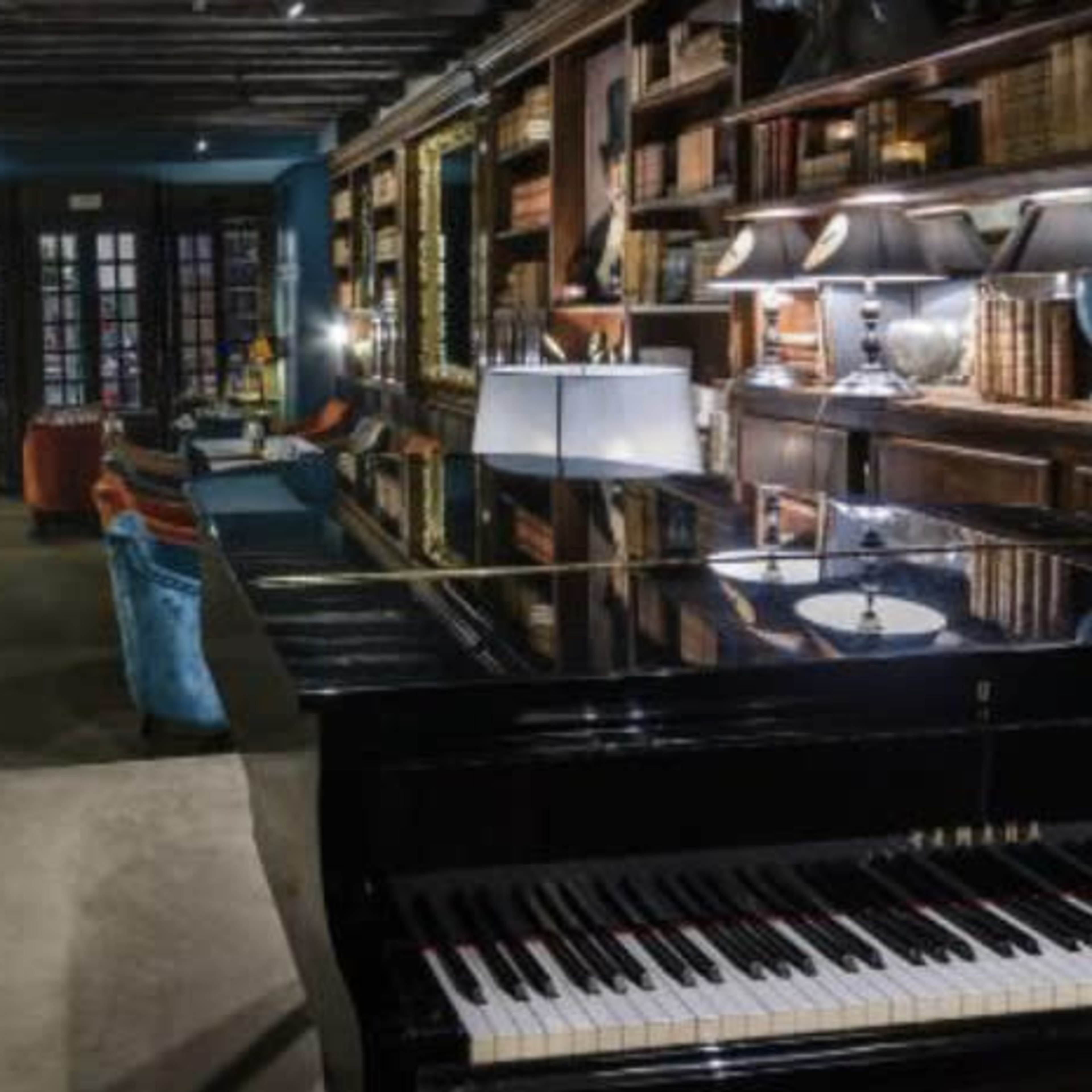 An elegant dining area with wooden shelves filled with books, a grand piano in the foreground, and tables set for dining in a softly lit atmosphere.