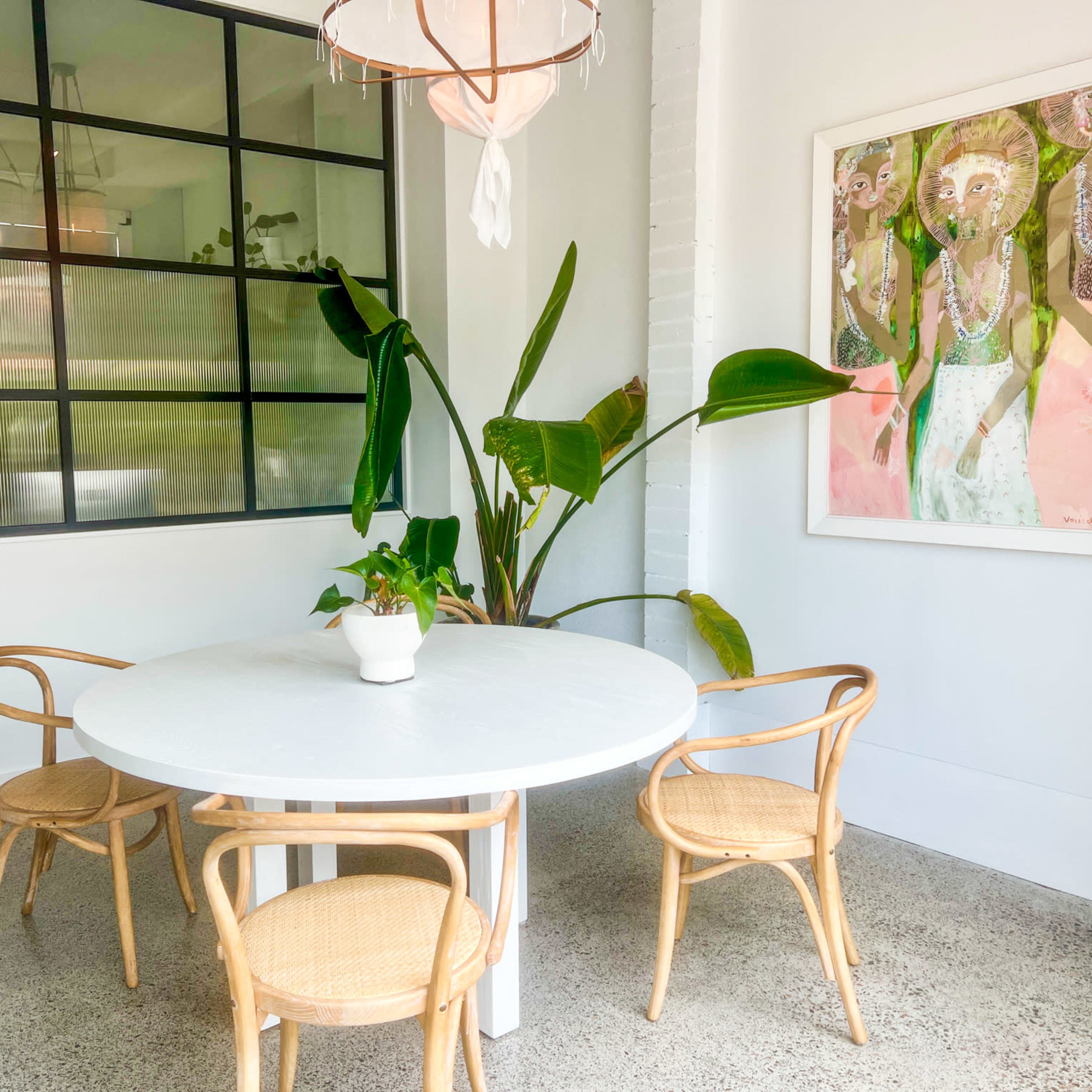 A round white table with four woven chairs is positioned in a bright room featuring a large window and a potted plant.