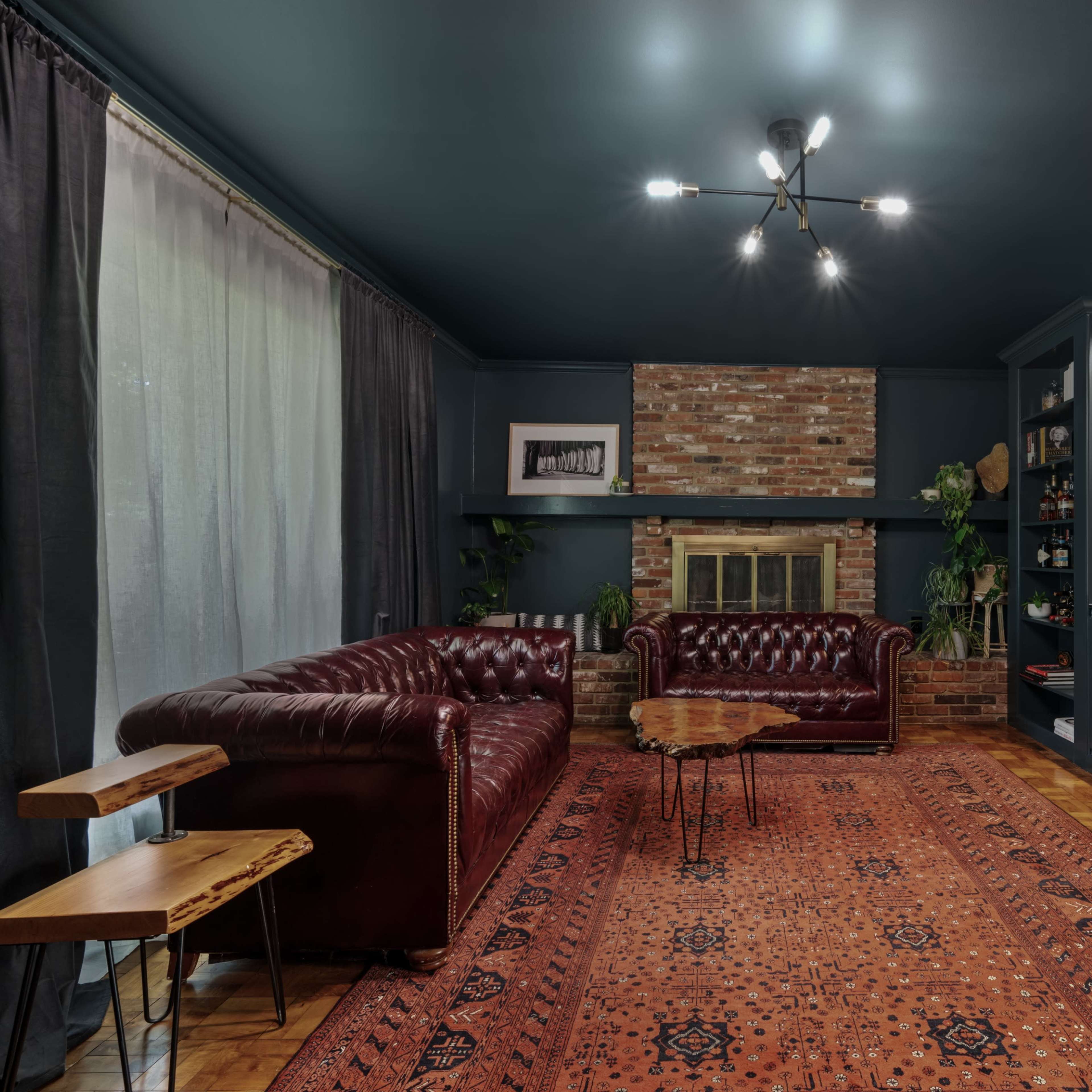 A cozy living room featuring a dark blue ceiling, a brick fireplace, a burgundy leather sofa, and a patterned area rug.