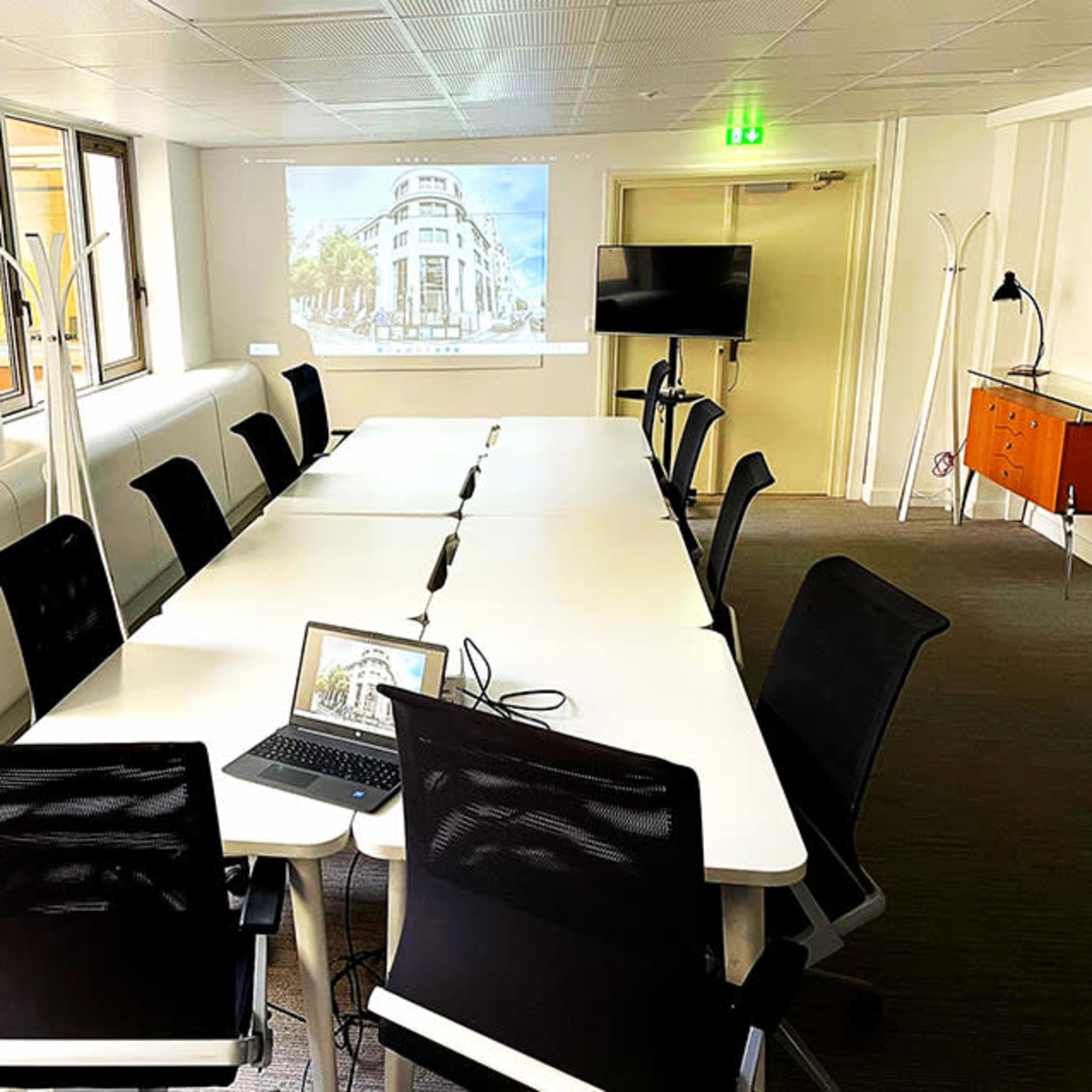 A modern conference room features a long table surrounded by black chairs, with a laptop on the table and a projector displays an image on the wall.