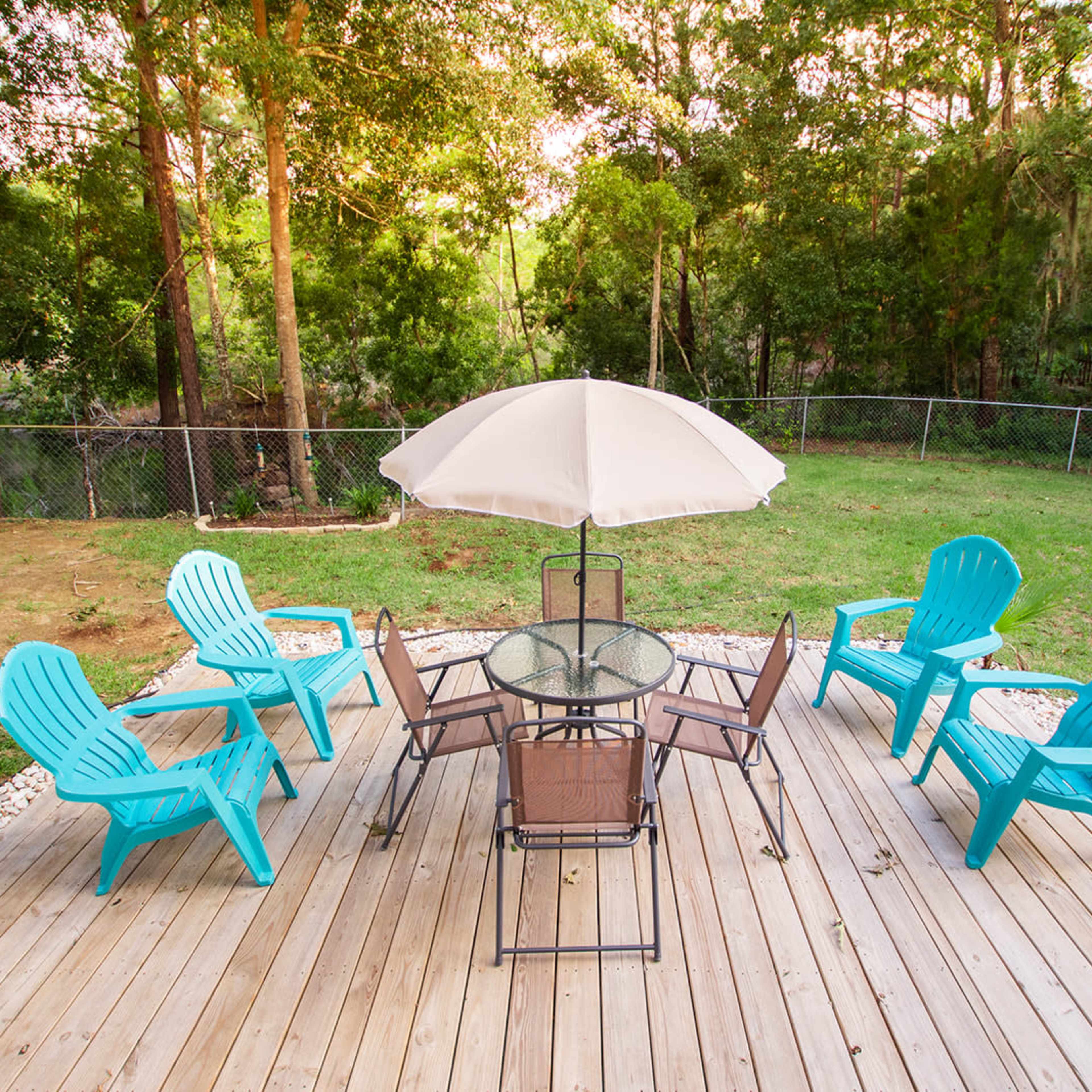 A wooden deck features a circular glass table surrounded by six blue adirondack chairs and a large umbrella, set against a backdrop of trees and a grassy area.