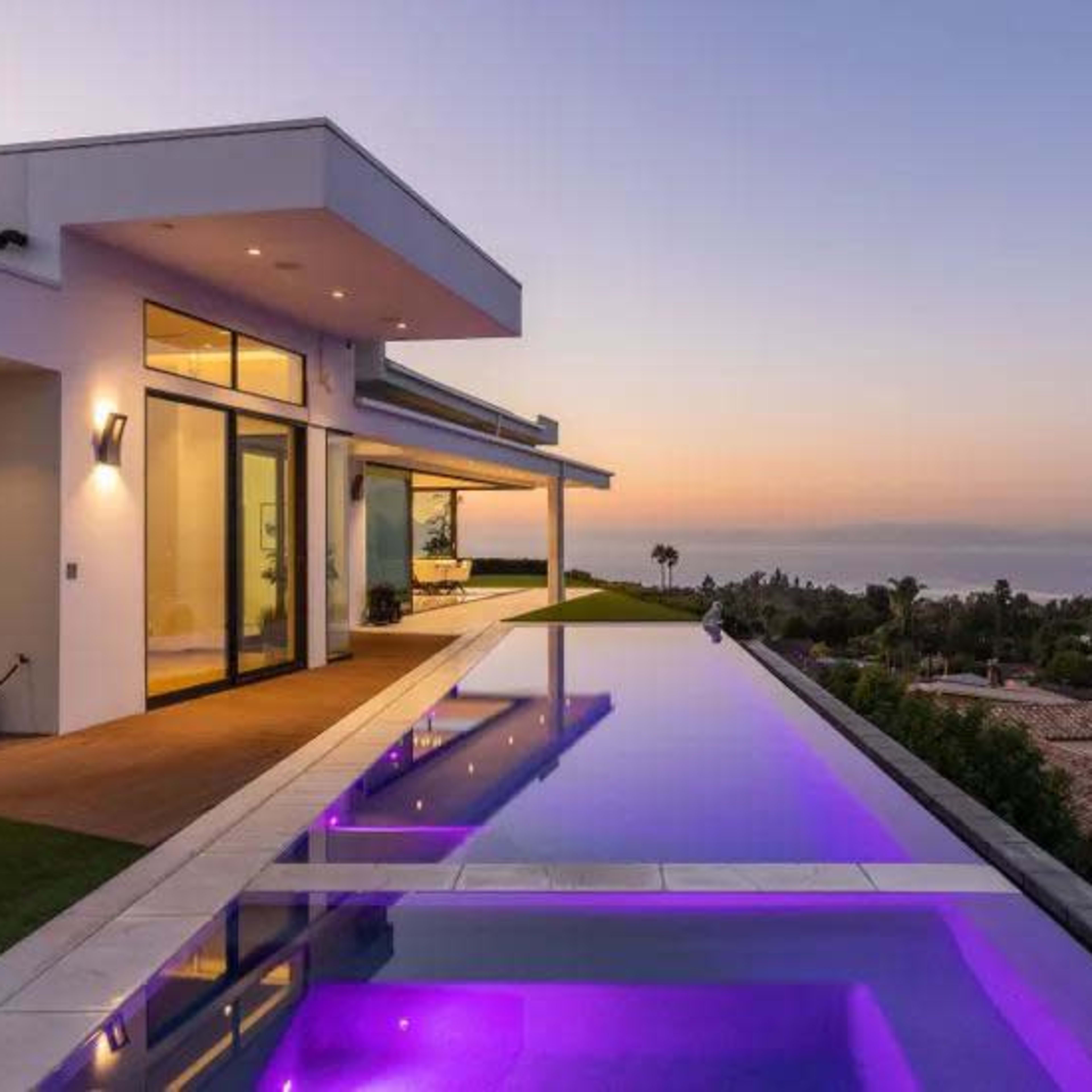 A modern house with large glass windows overlooks a pool illuminated with purple lights at sunset.