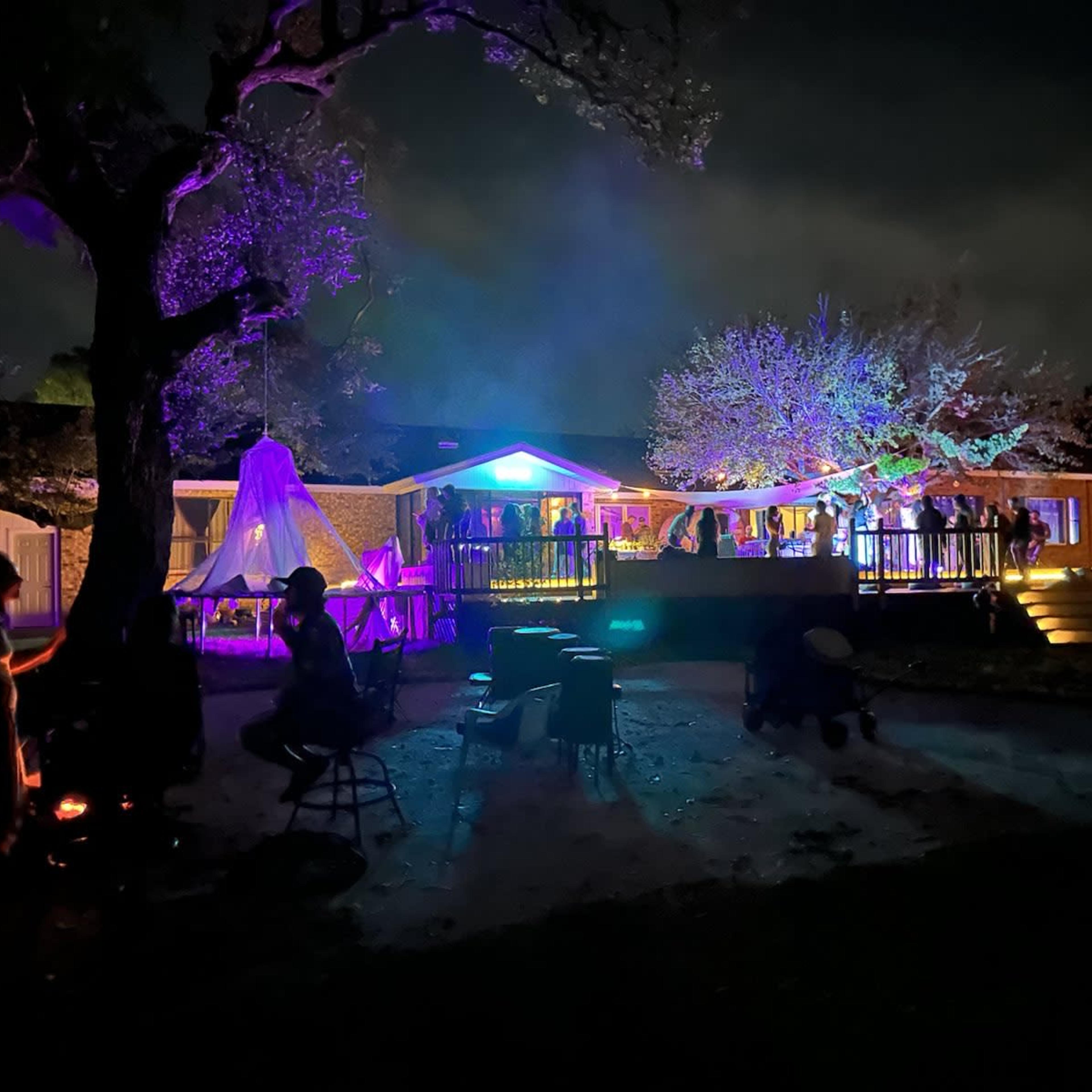 A gathering takes place outdoors at night, illuminated by colorful lights with people socializing near a bonfire and a house in the background.