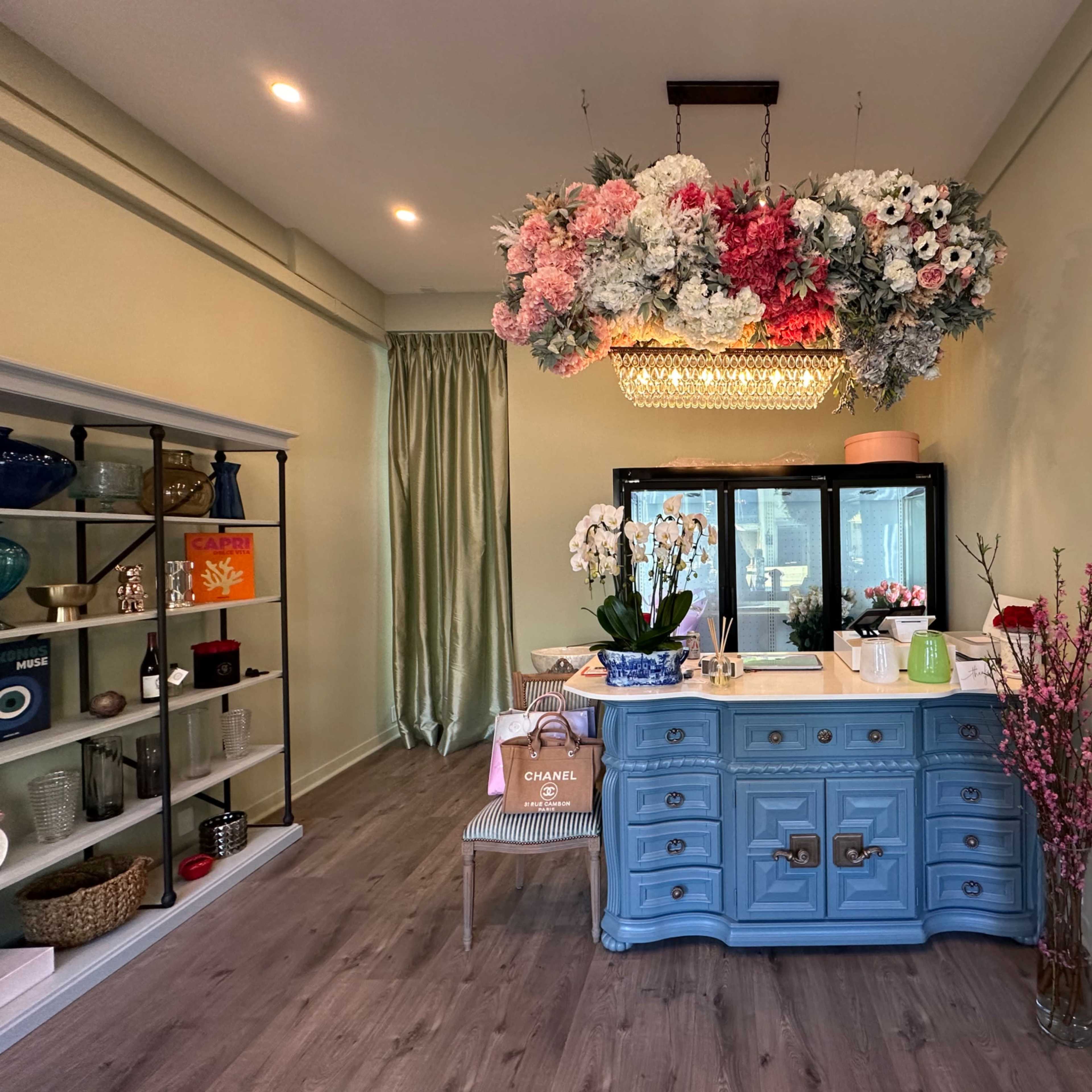 A light blue dresser with decorative flowers overhead stands in a softly lit room featuring a mirror and shelves filled with various items.