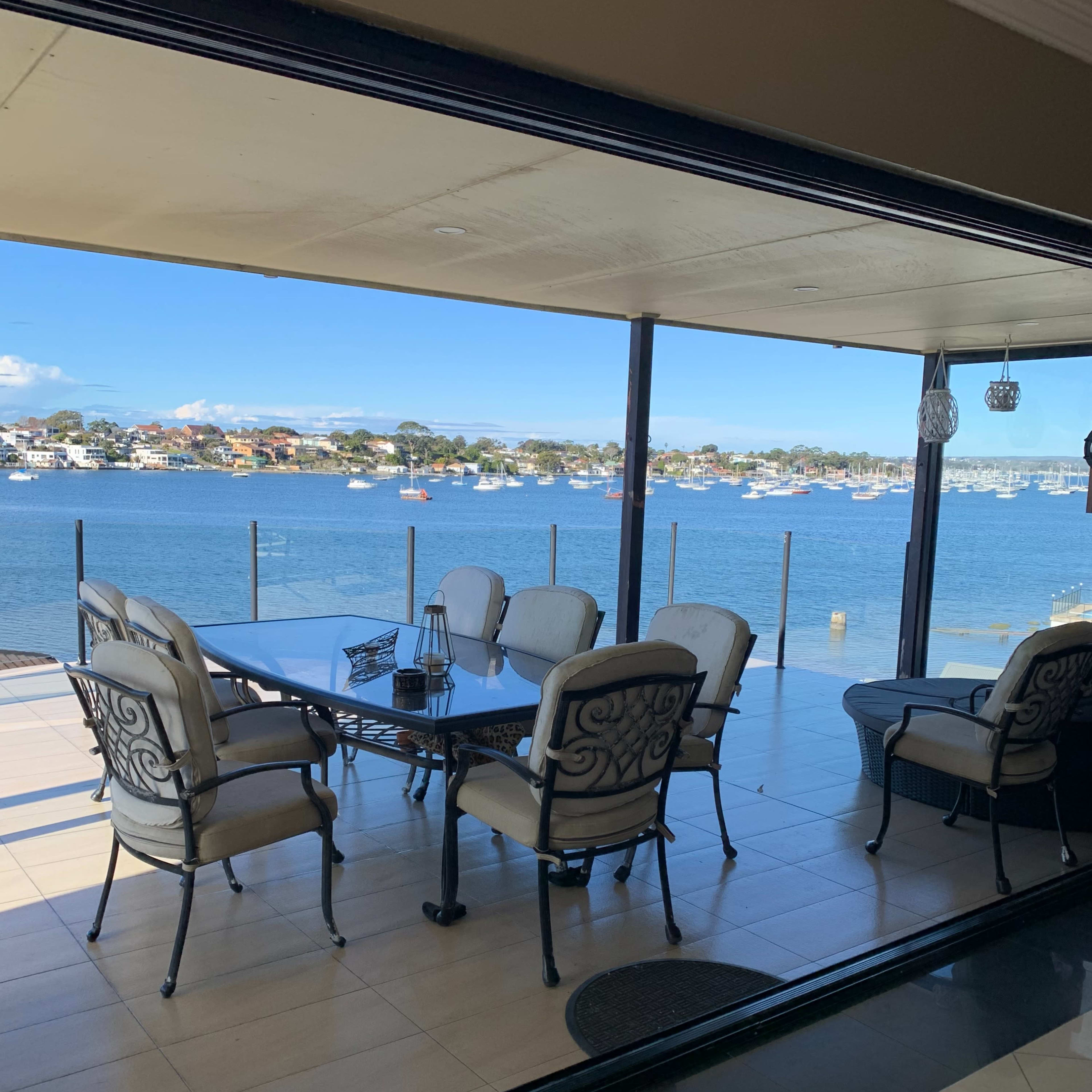 A covered patio with a table and chairs overlooks a waterfront view with boats and houses in the background.