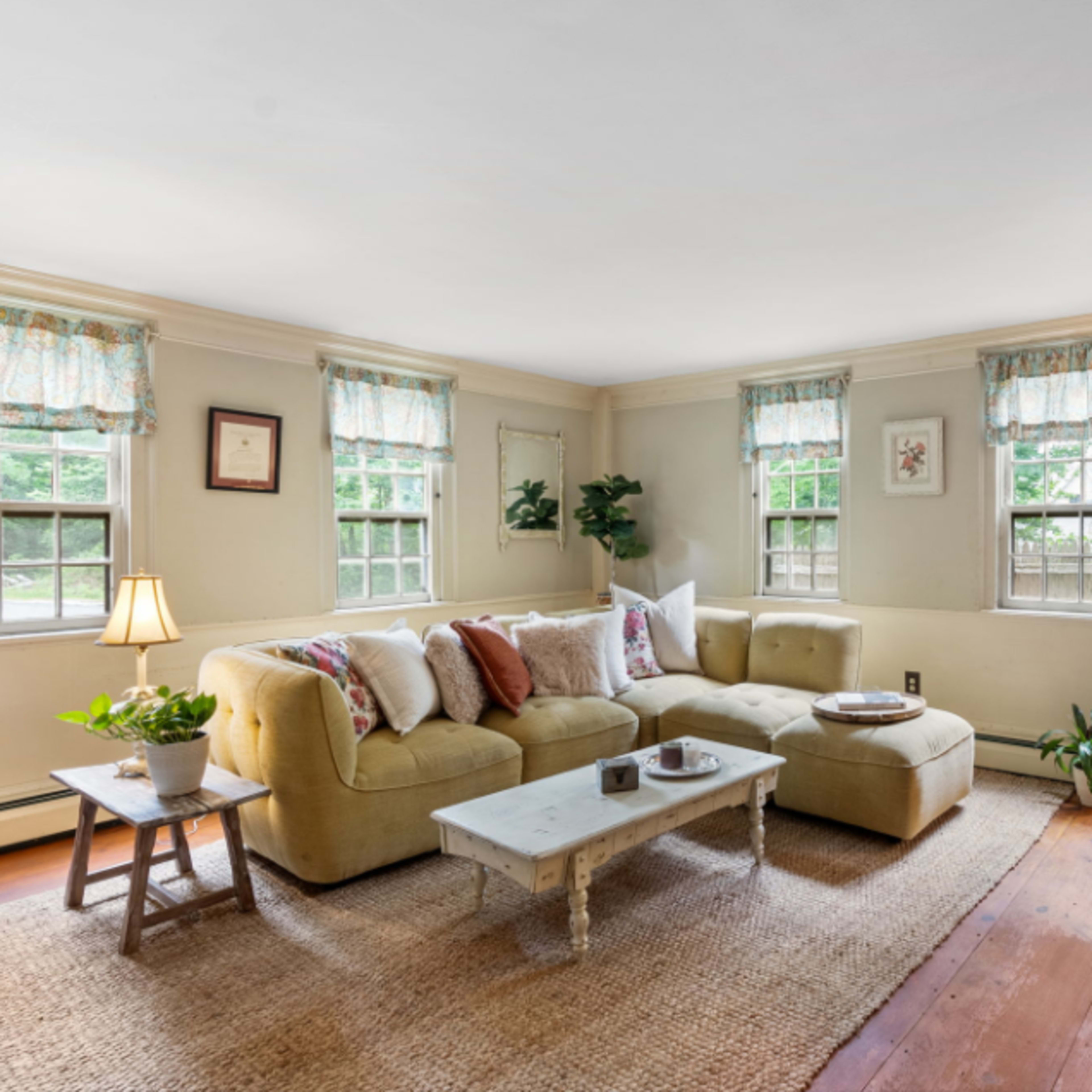 A bright living room features a light-colored sectional sofa, a wooden coffee table, and several windows allowing natural light to stream in.