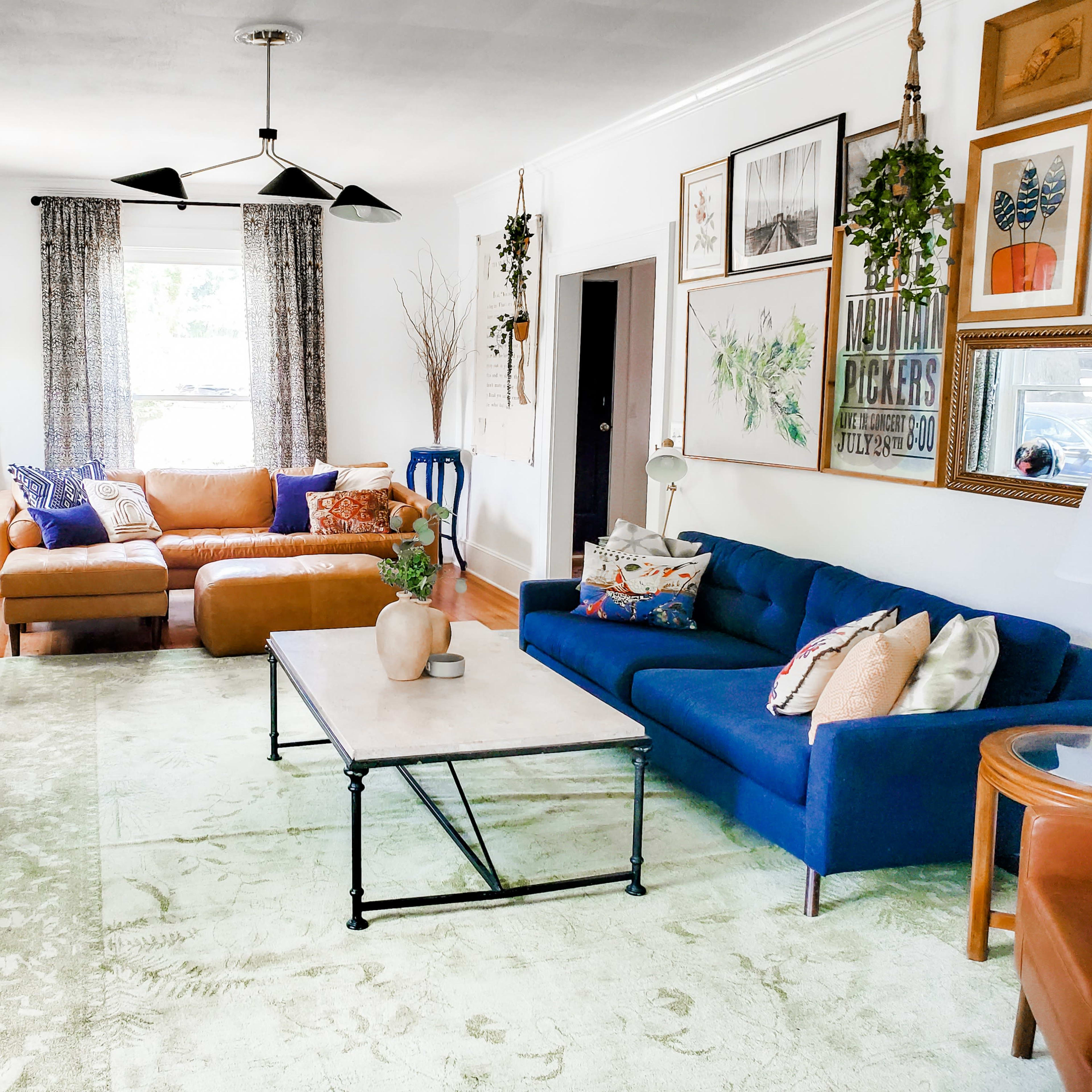 The living room features a blue sofa, a brown sectional, a coffee table, and various plants, framed artwork, and decorative items arranged around a light-colored rug.
