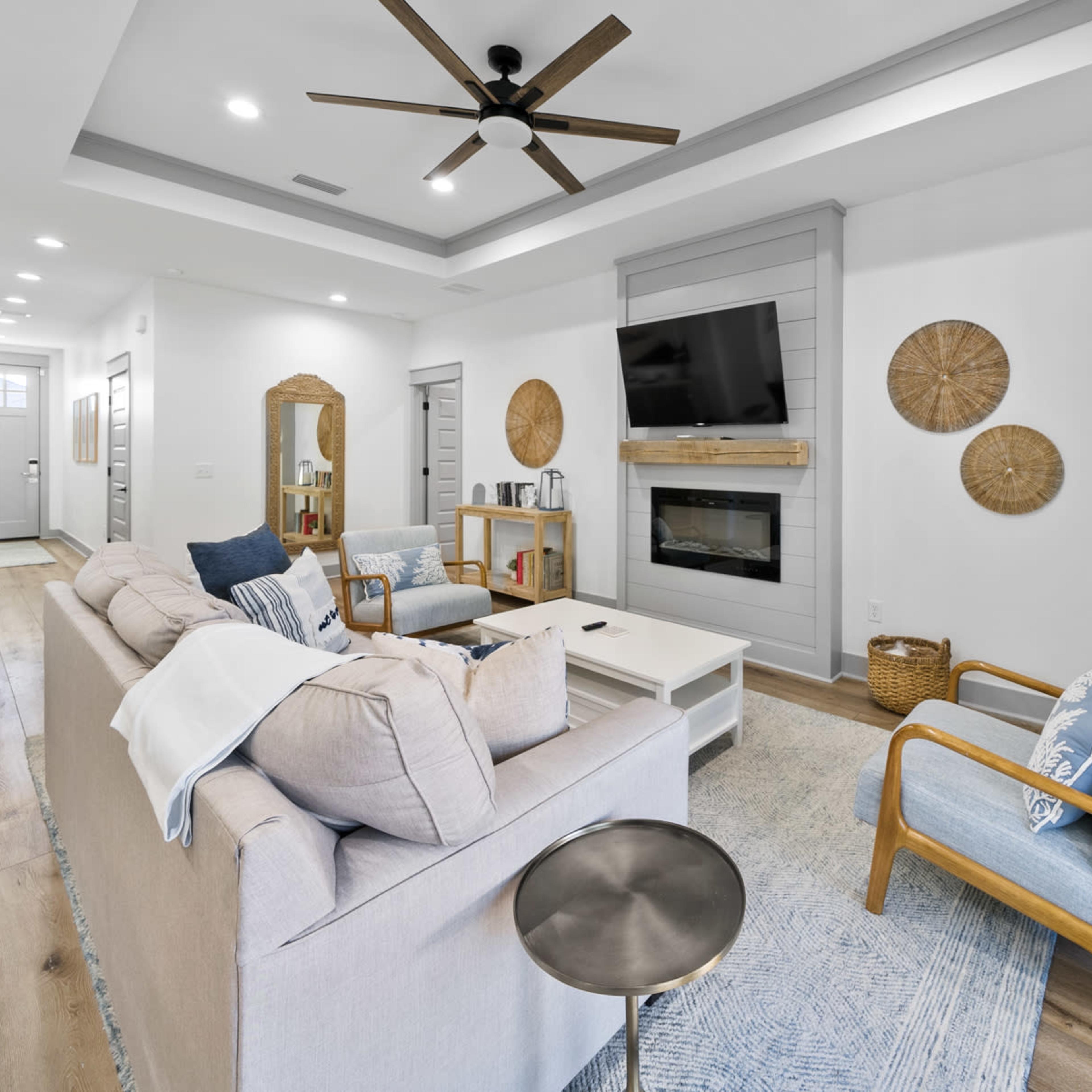 A modern living room with a light-colored sofa, a wooden coffee table, and a flat-screen TV mounted on a wall, complemented by decorative wall hangings and a plant.