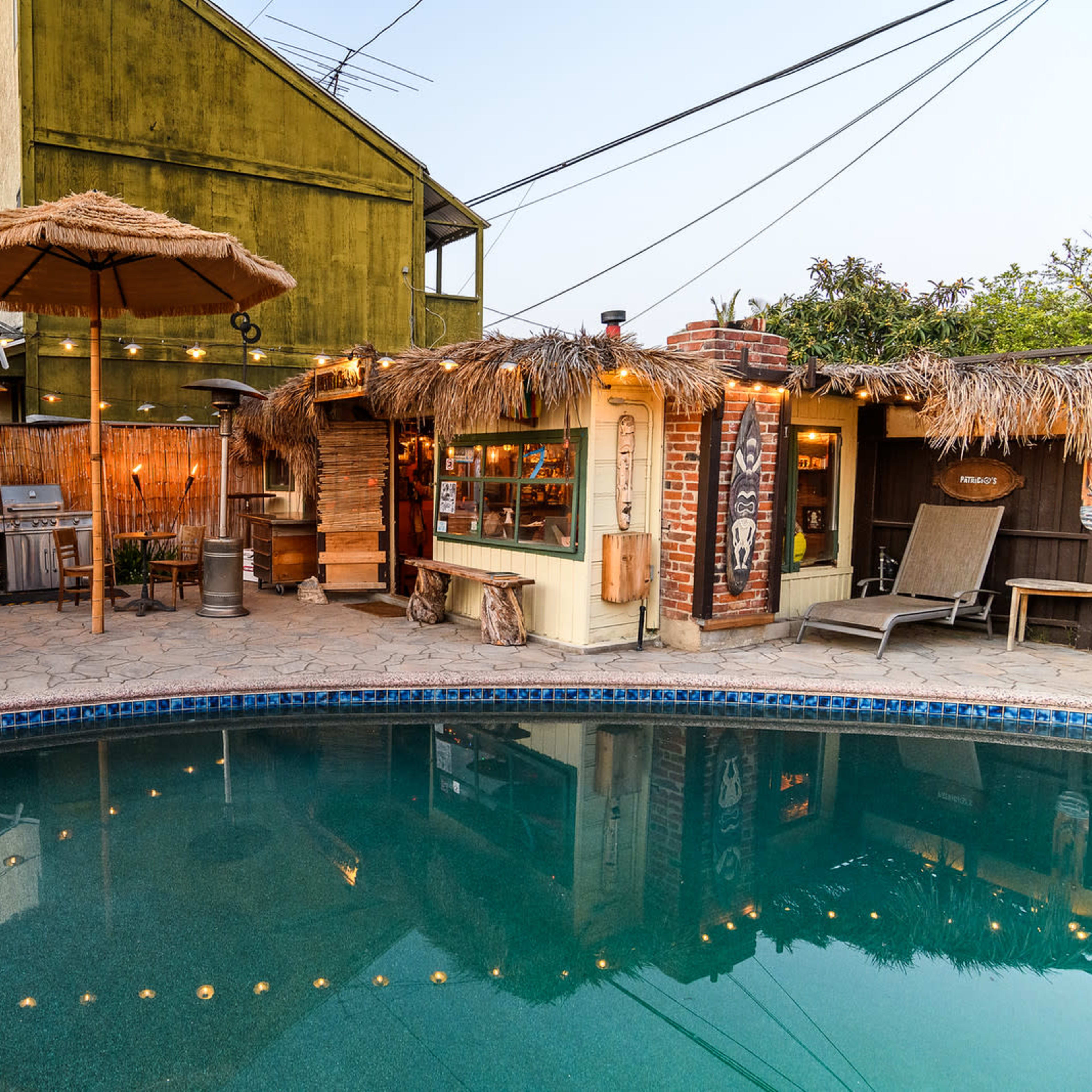 A backyard scene featuring a swimming pool surrounded by a stone patio, a thatched umbrella, and string lights illuminating a bar area with seating.