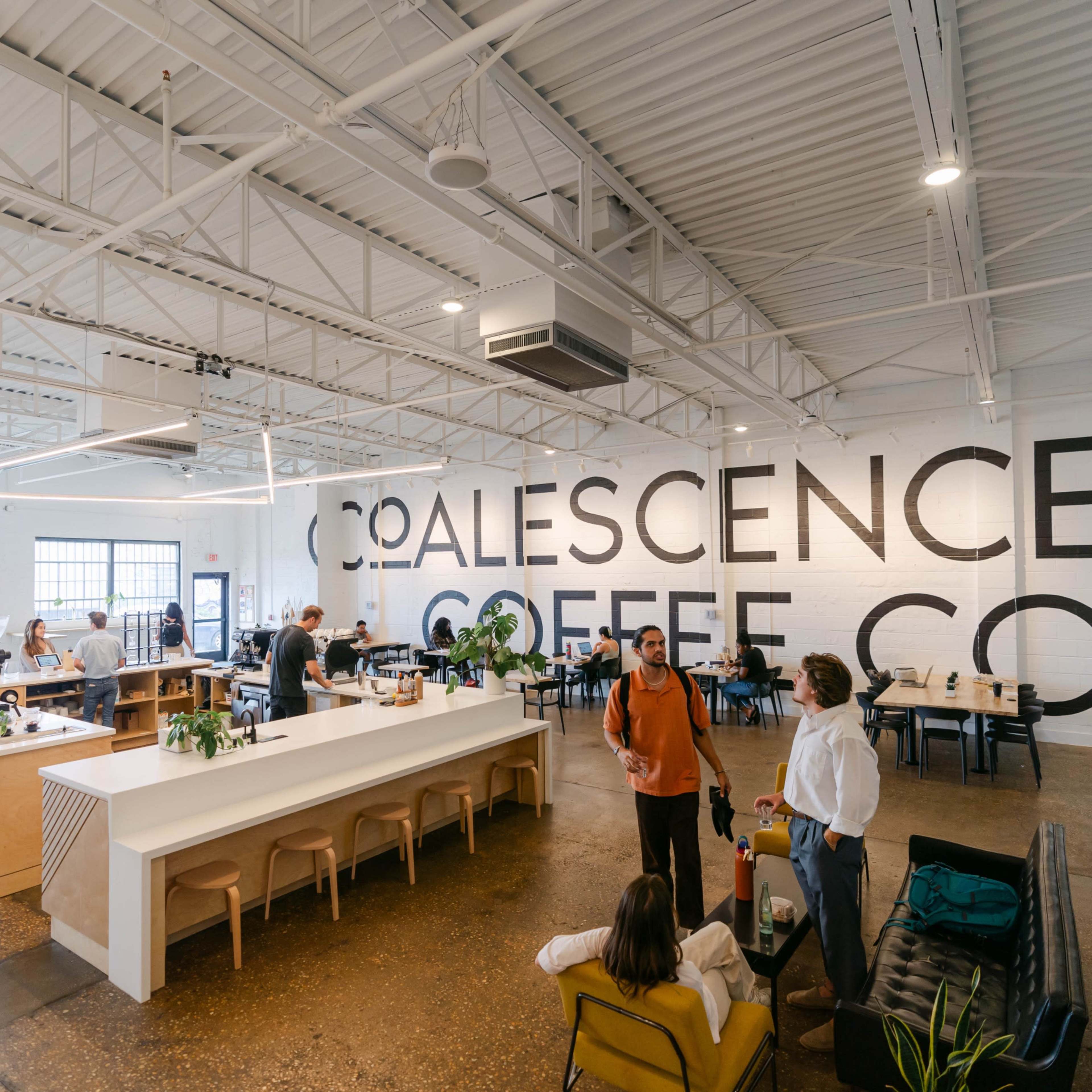 A modern coffee shop with a spacious interior featuring a large counter, seating areas, and a prominent wall displaying the name "Coalescence Coffee Co."