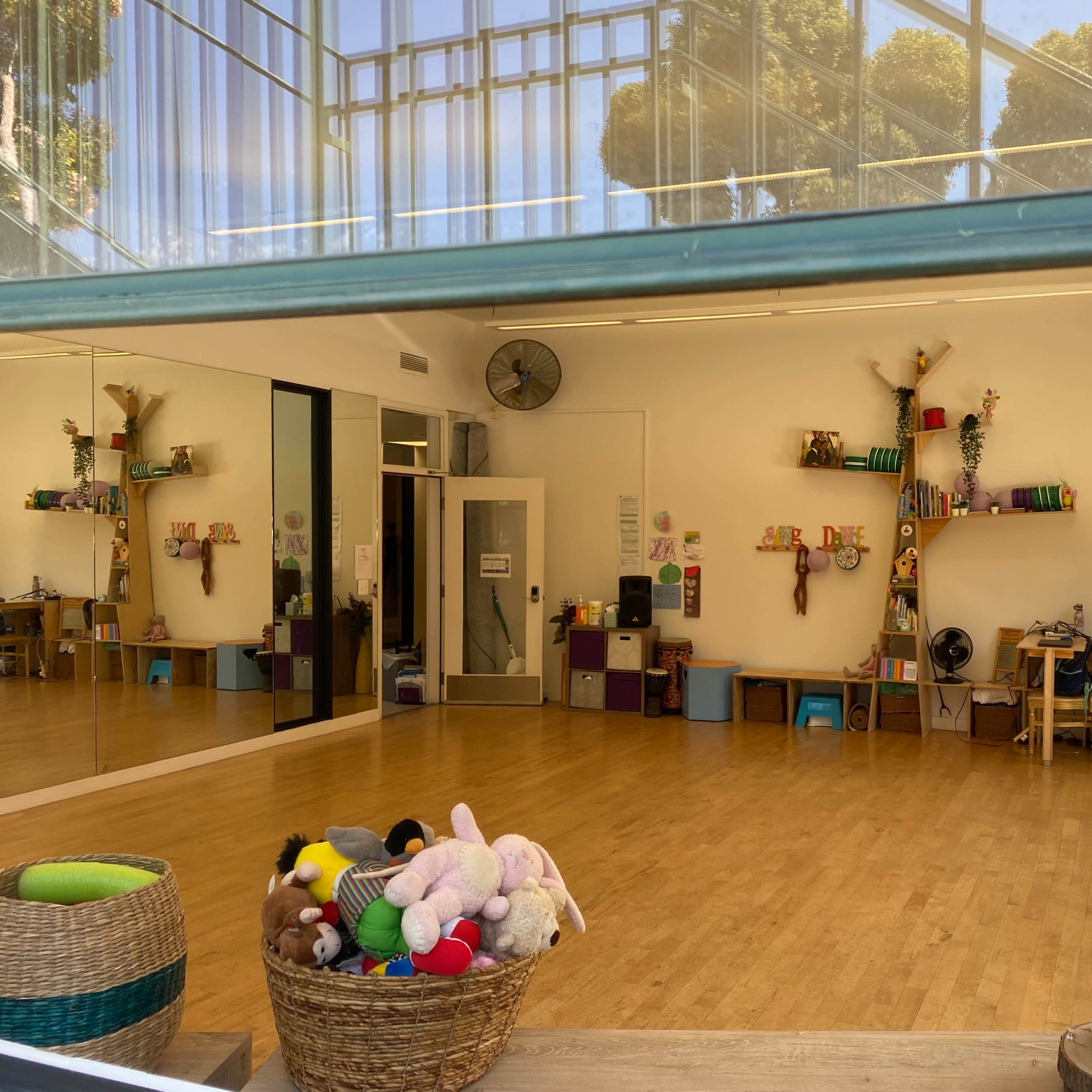 A bright, spacious room with wooden flooring, shelves decorated with colorful items, and a basket filled with stuffed animals in the foreground.