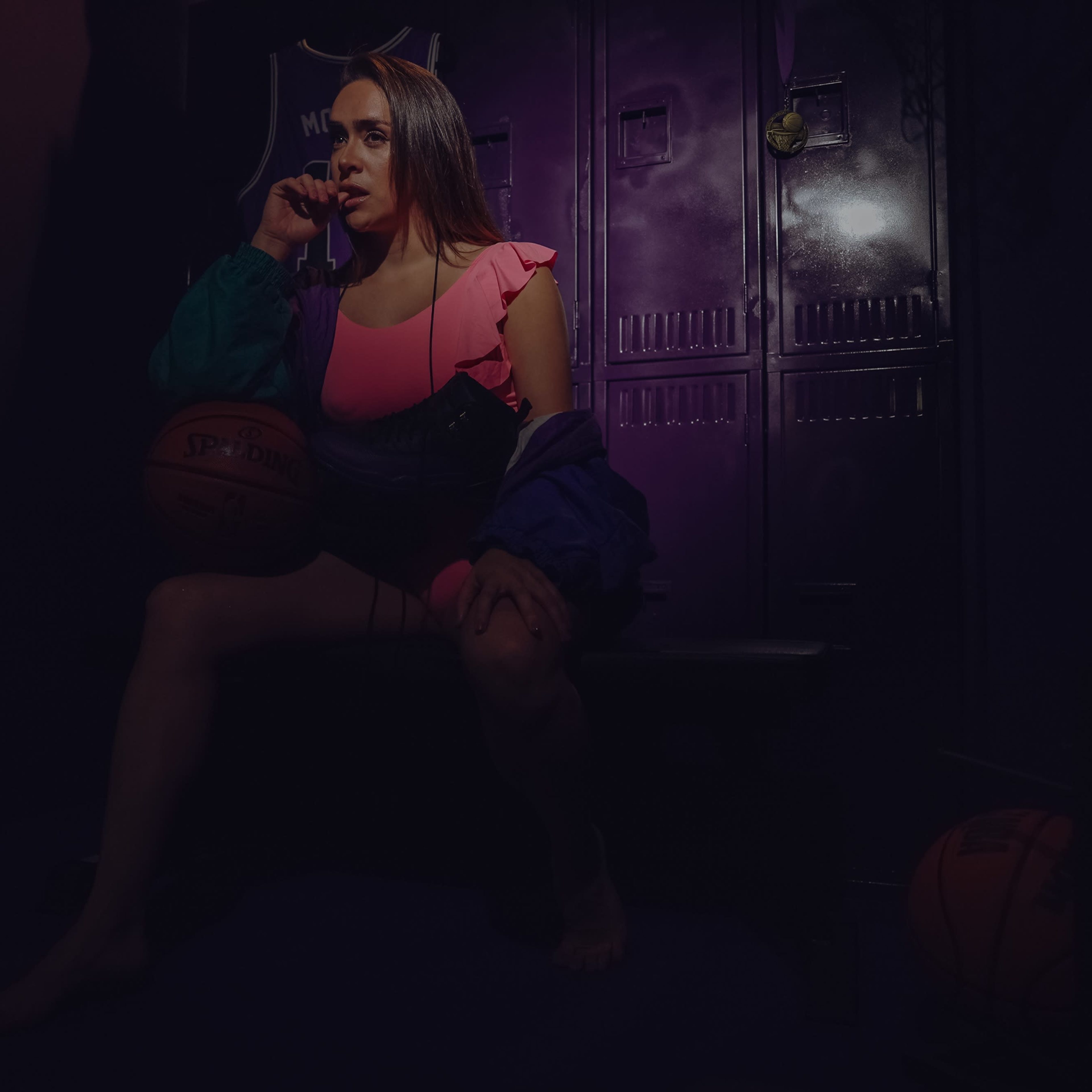 A woman in a pink outfit and colorful jacket sits on a bench in front of purple lockers, holding a basketball.
