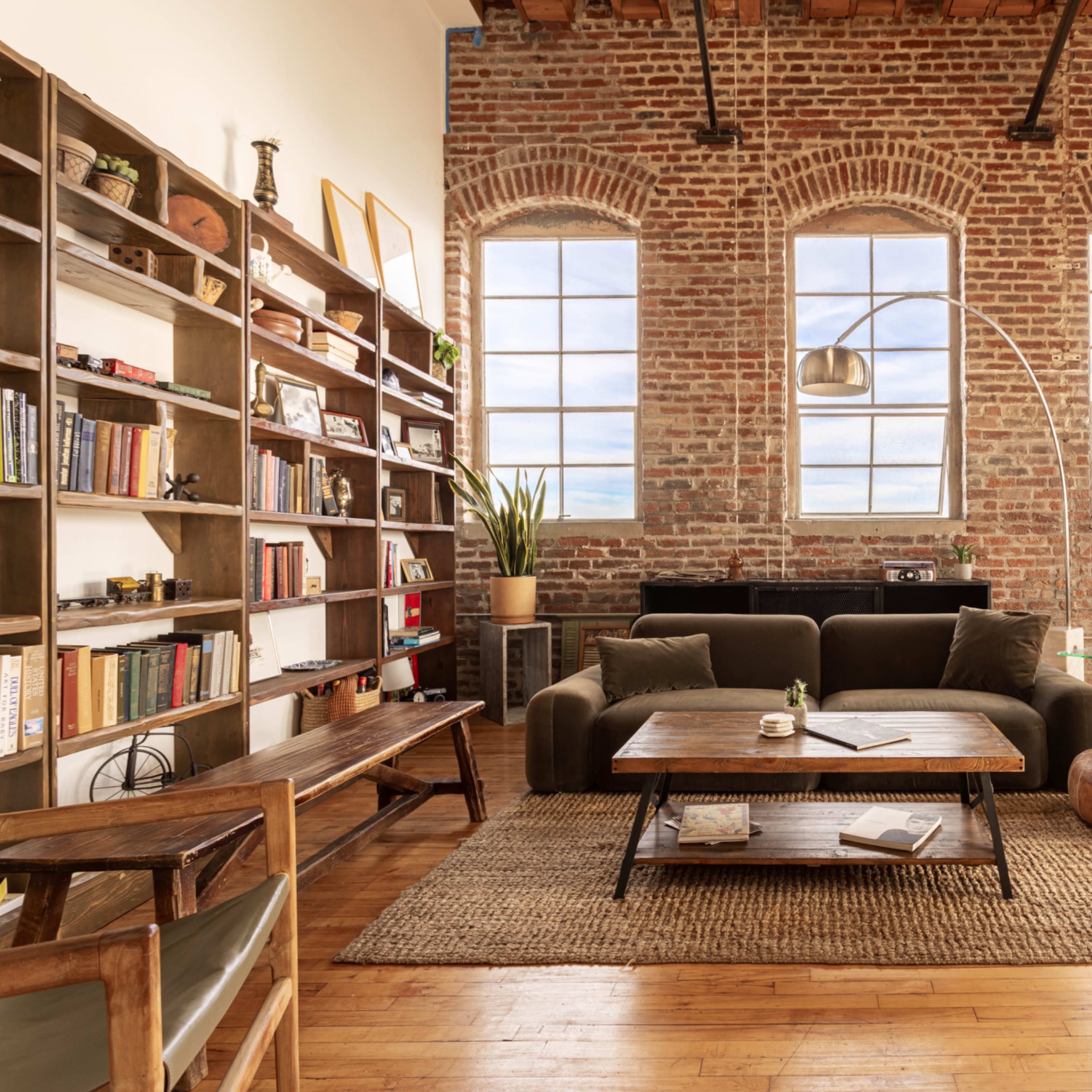 A spacious living area features a mix of modern furniture and a bookshelf against a wall of exposed brick, with large windows allowing natural light to fill the room.
