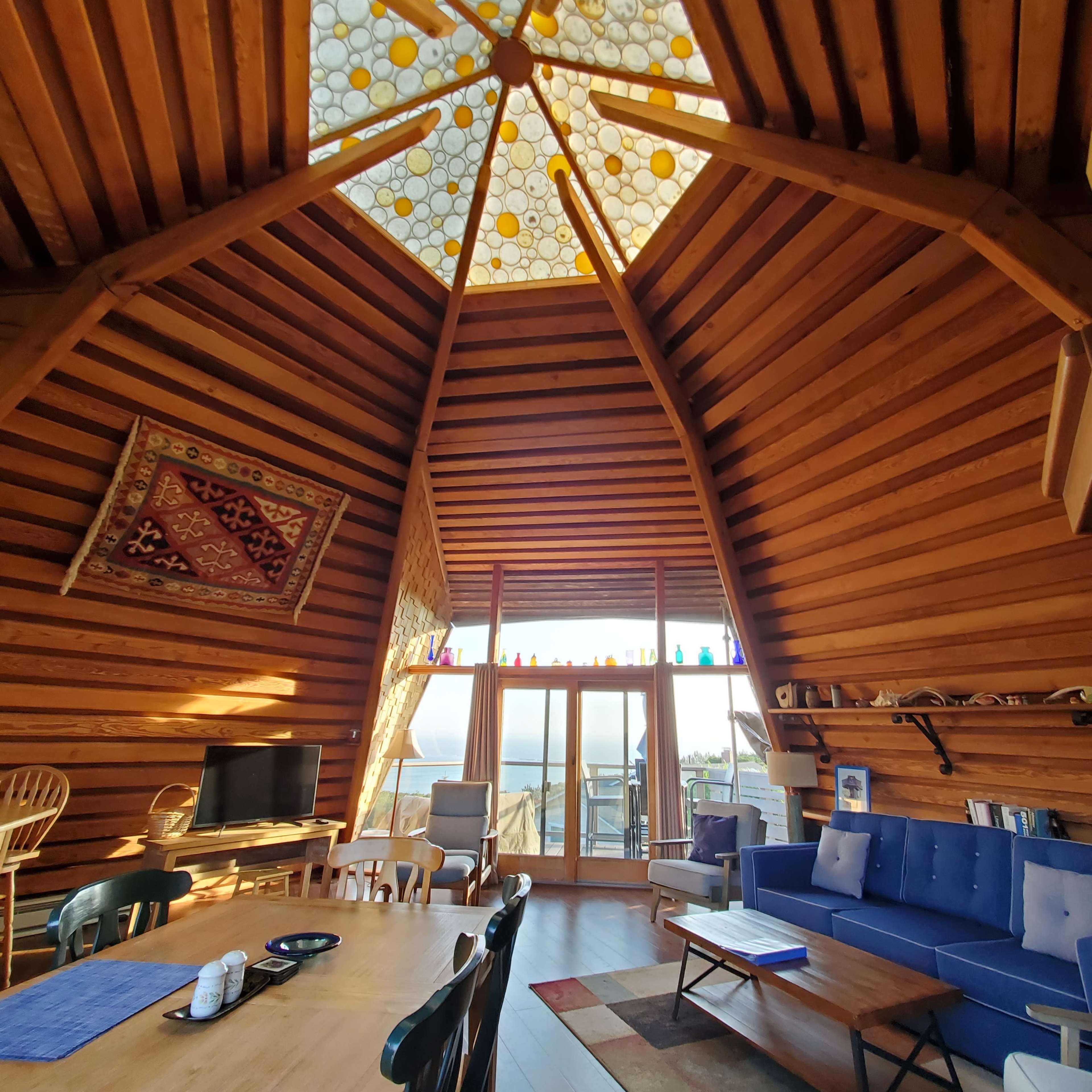The interior of a wooden cabin features a high, angled ceiling with a glass skylight and a spacious living area furnished with a table, chairs, and a couch.