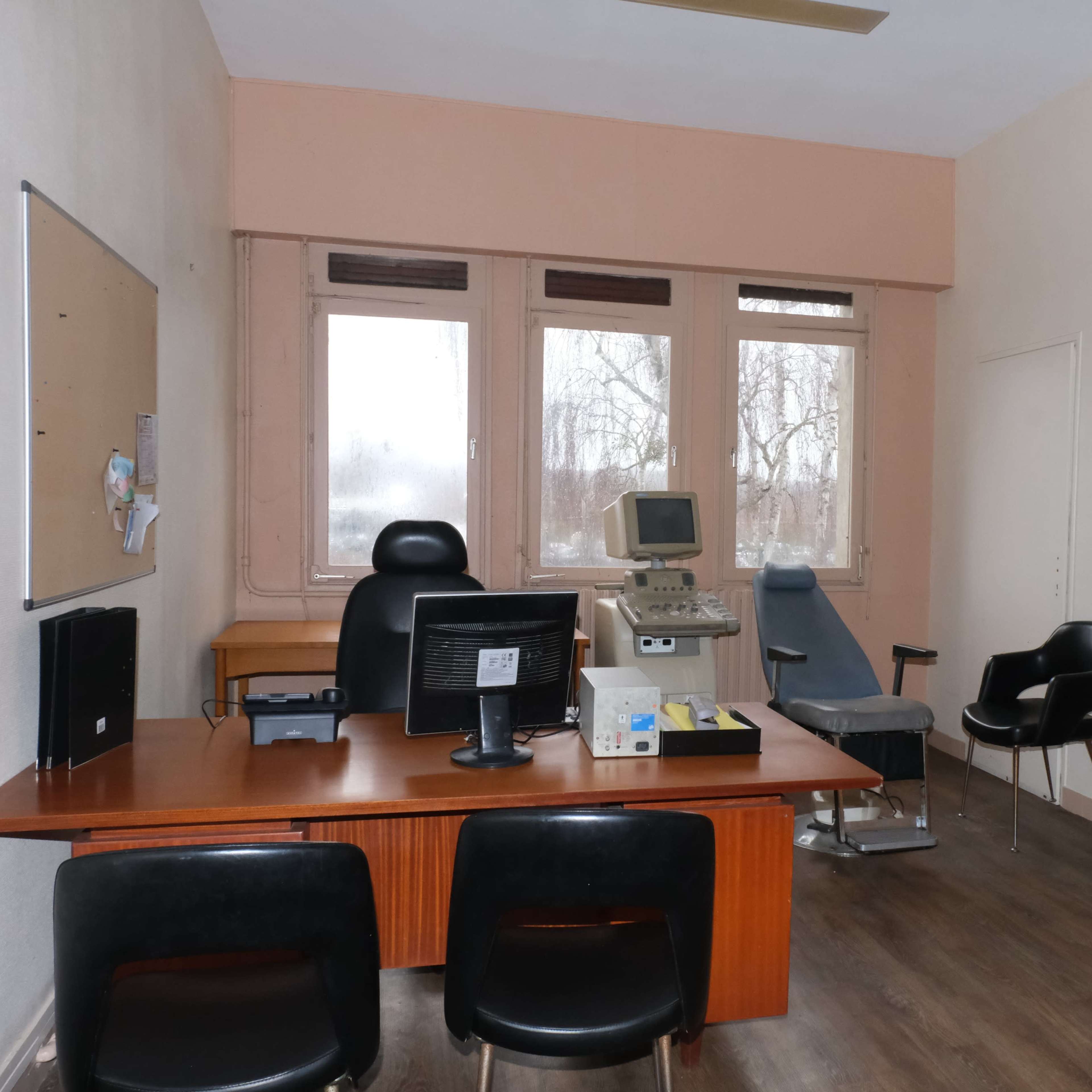 A small medical office featuring a desk with a computer, an examination chair, and a wooden cabinet.