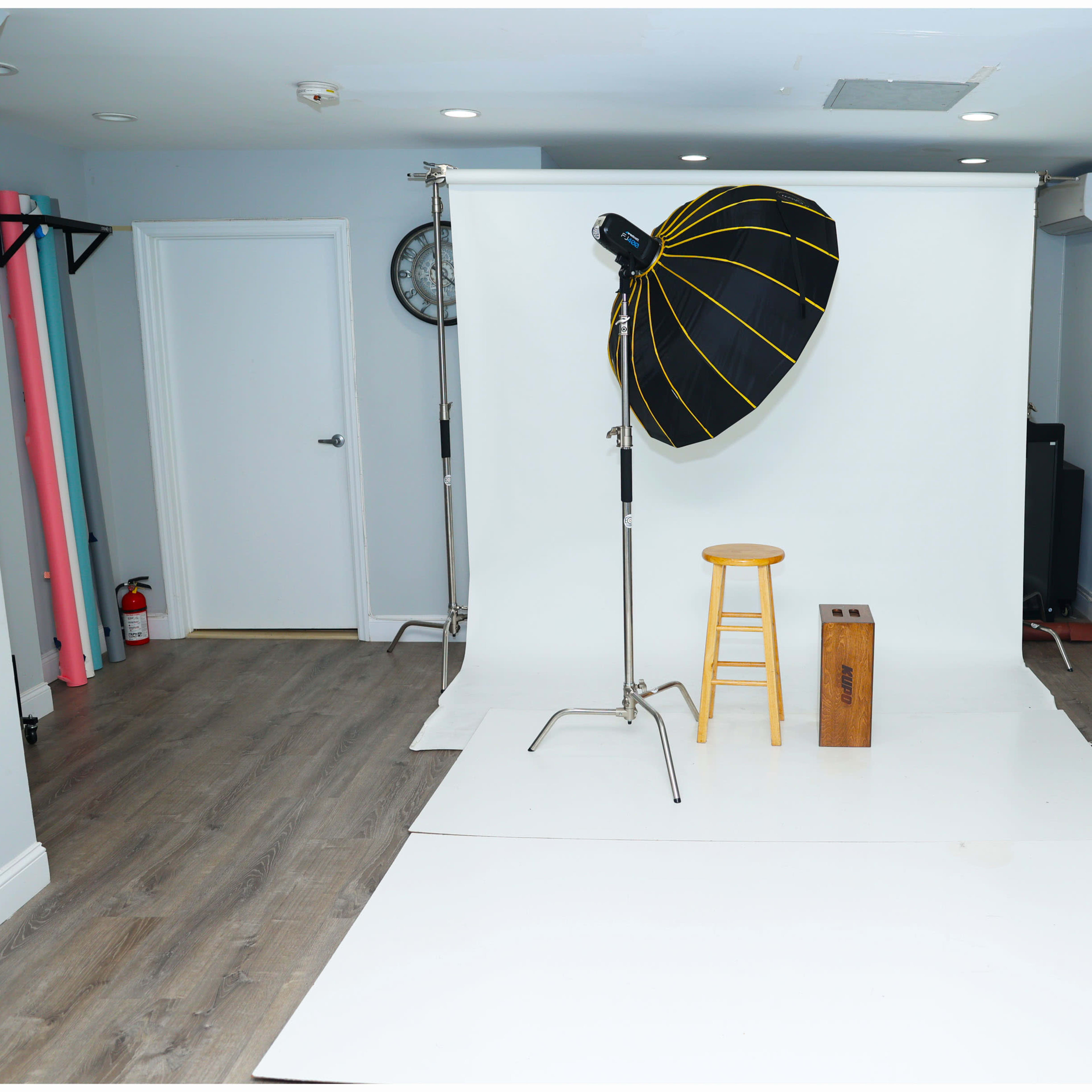 A photography studio with a white backdrop, a large softbox light, a wooden stool, and a small wooden box.