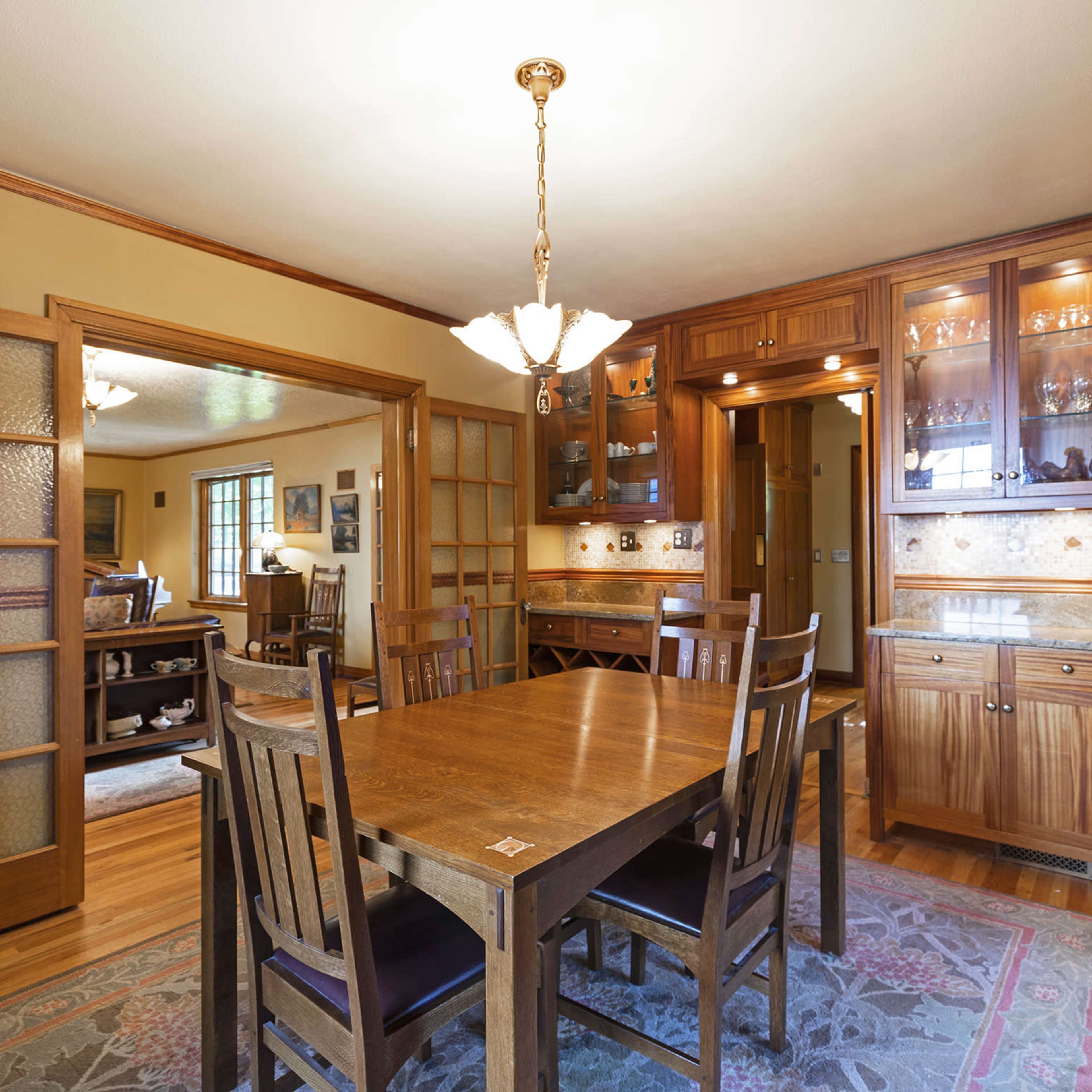 A wooden dining room with a large table and a built-in cabinet displaying glassware, featuring warm lighting and hardwood flooring.