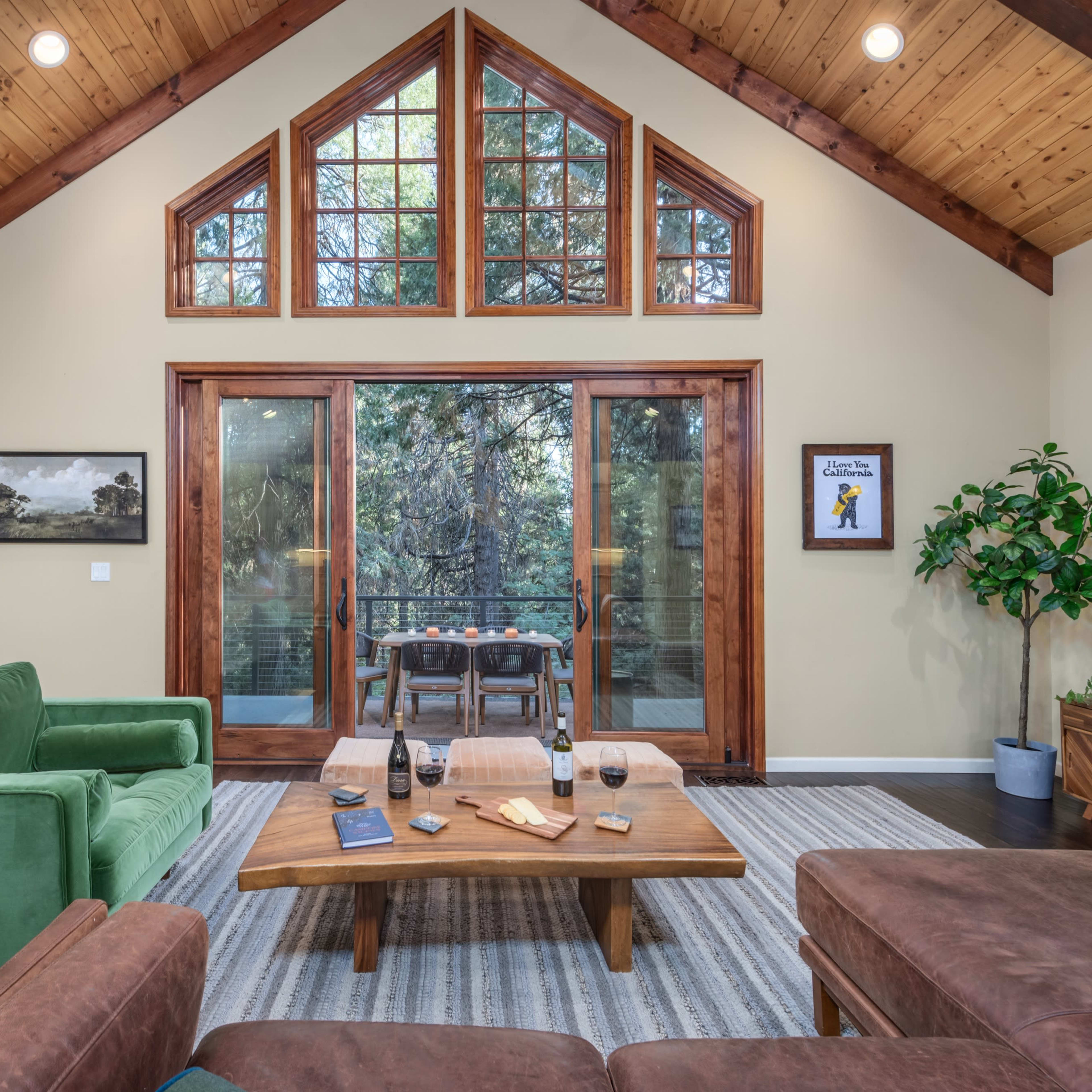 A cozy living room featuring a stone fireplace, green and brown couches, a wooden coffee table, and large windows with a view of greenery outside.